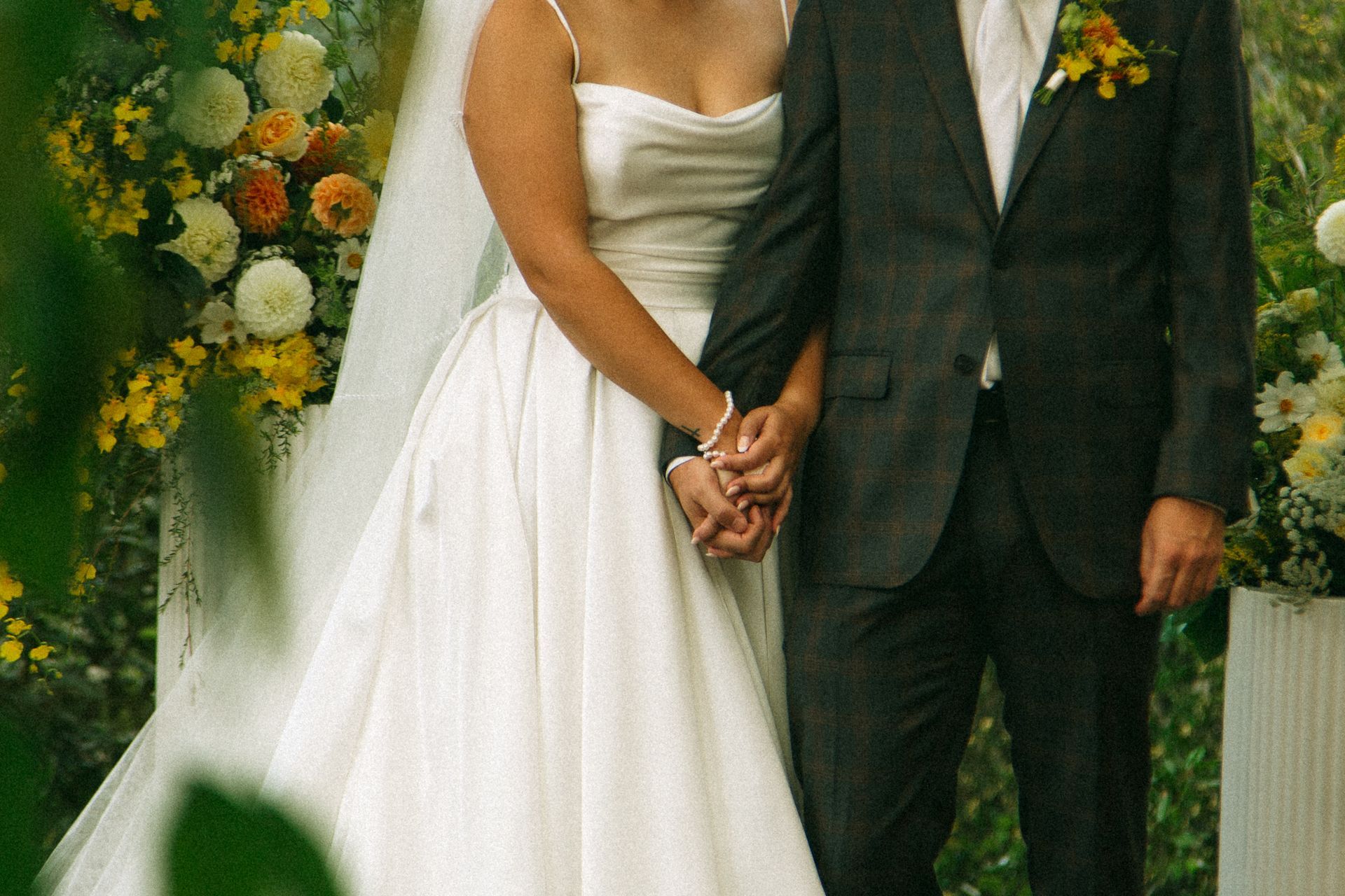 Bride and groom holding hands at outdoor wedding ceremony, flowers in background.