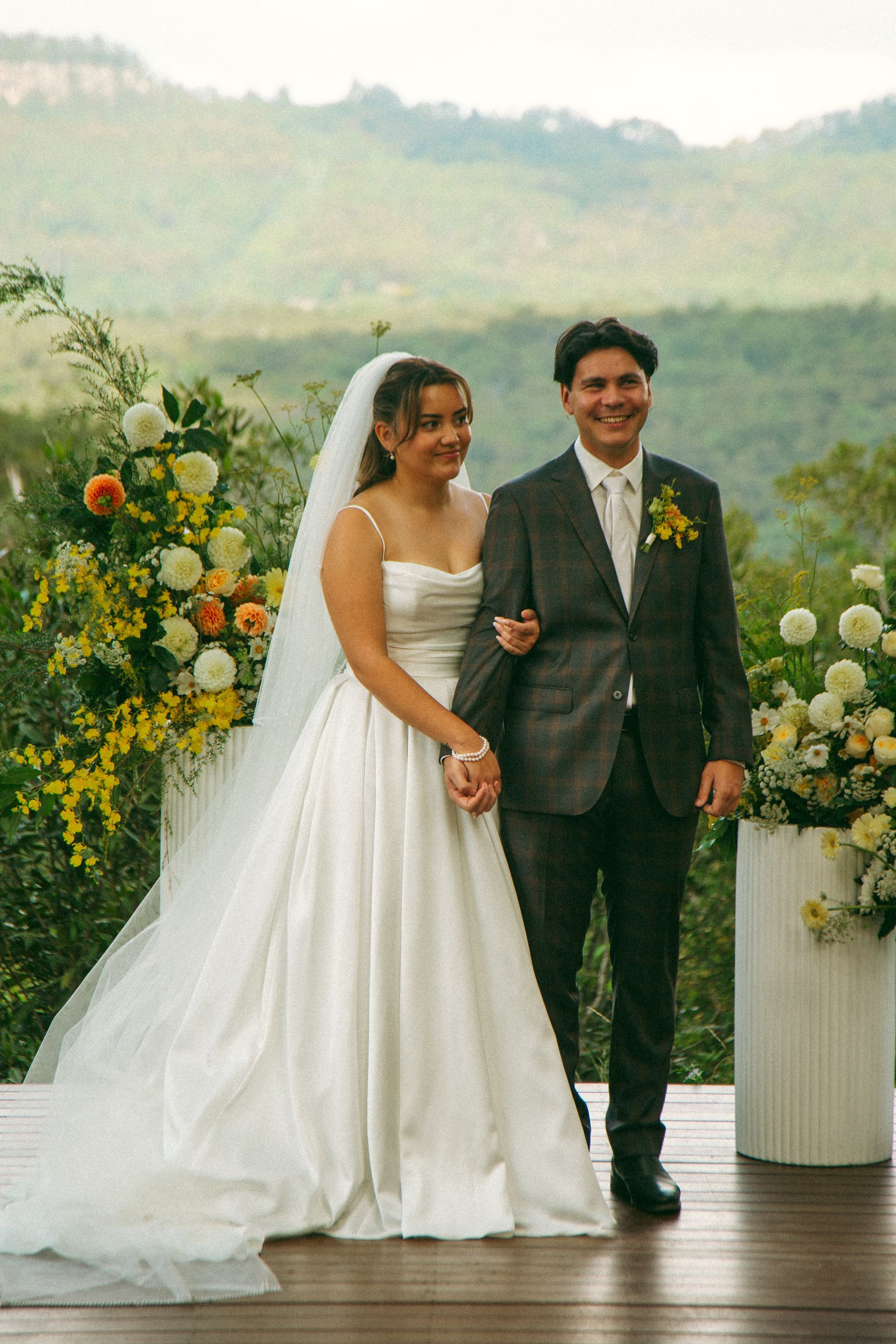 Bride and groom smiling, holding hands, after wedding ceremony. Outdoor setting, mountains in background.