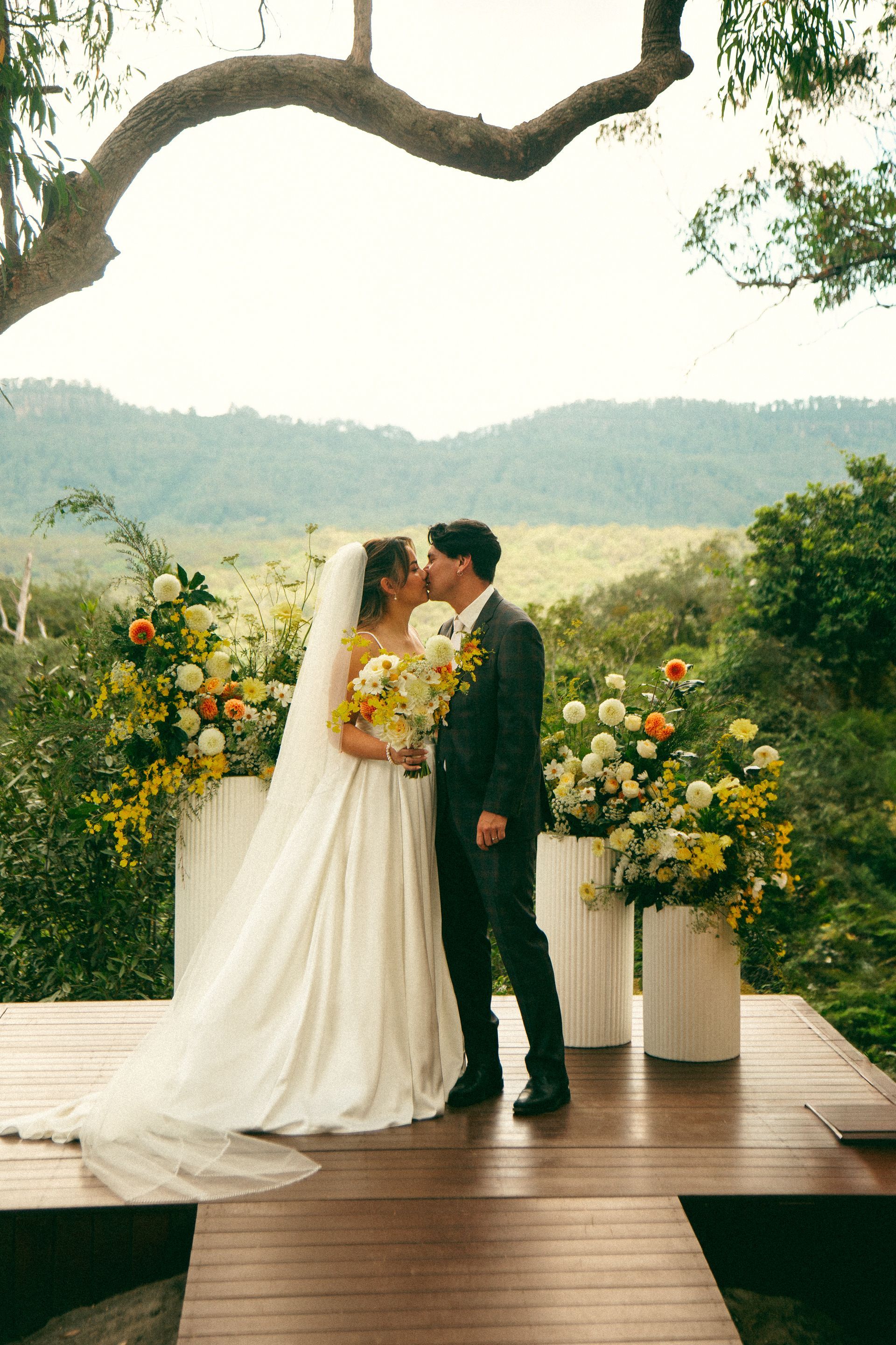 Bride and groom kissing at outdoor wedding ceremony with floral decorations and mountain backdrop.