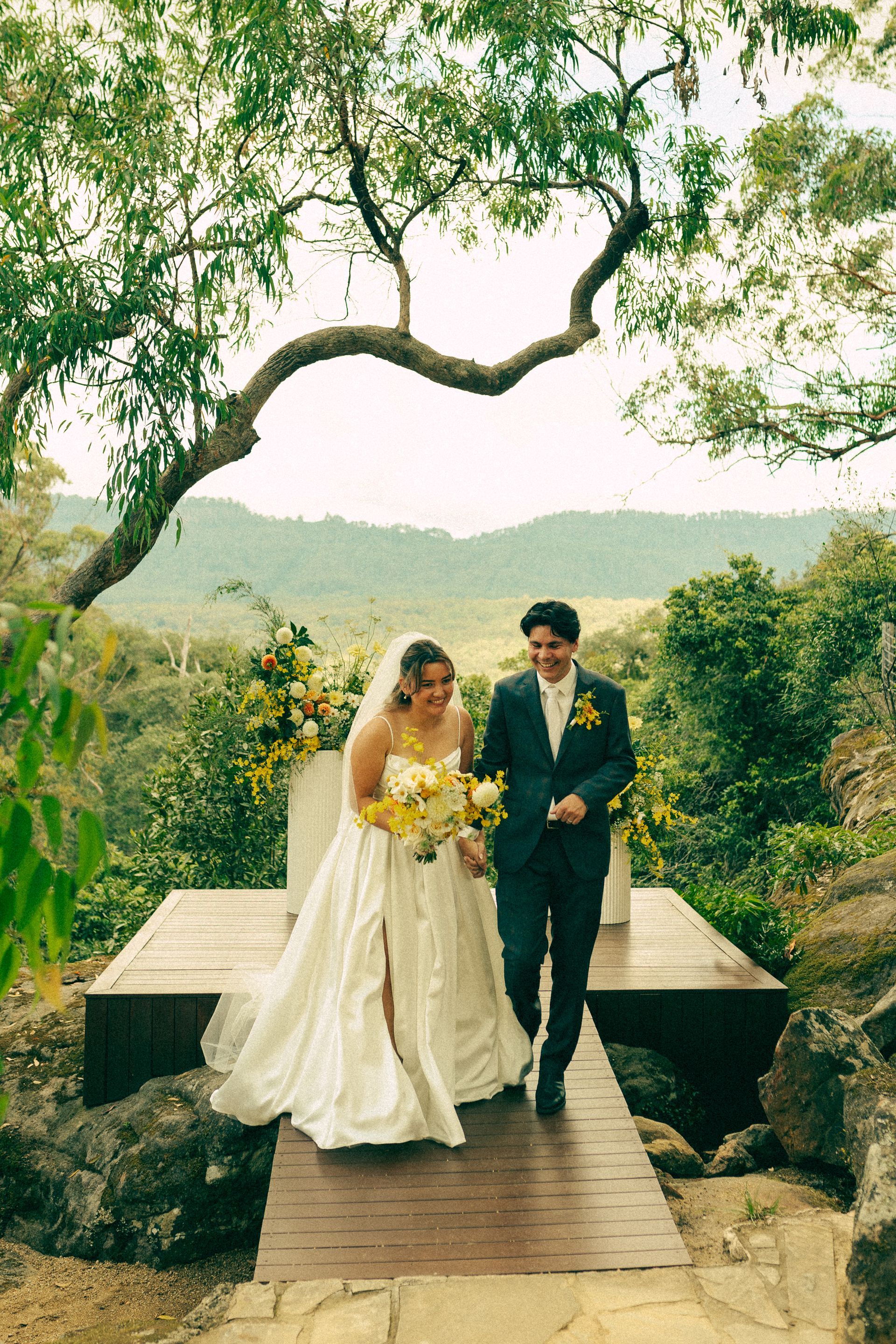 Bride and groom walking down wooden aisle, smiling, holding hands, lush greenery backdrop.