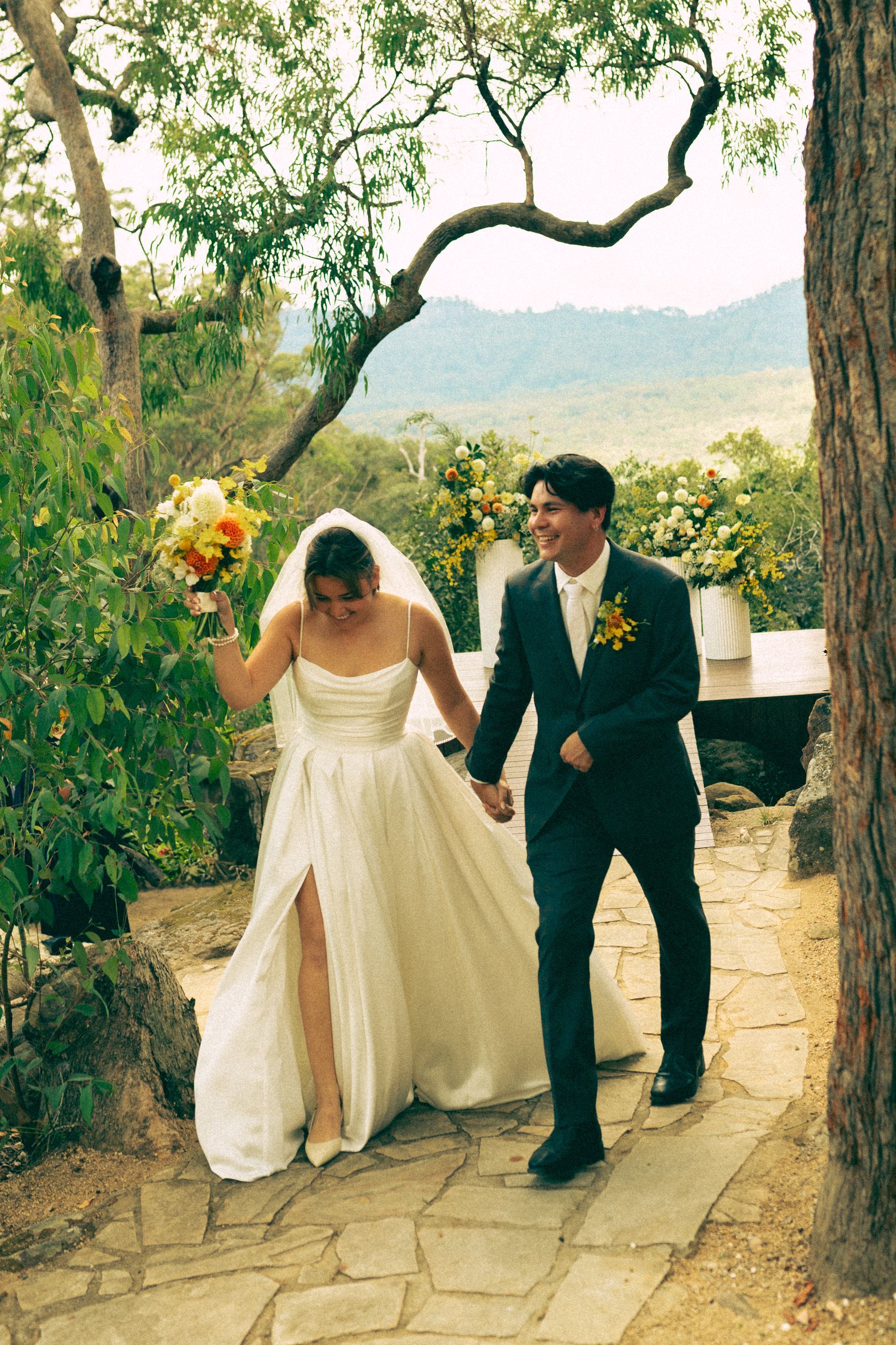 Bride and groom walk, holding hands, smiling. Bride in white dress, groom in suit. Outdoor setting.