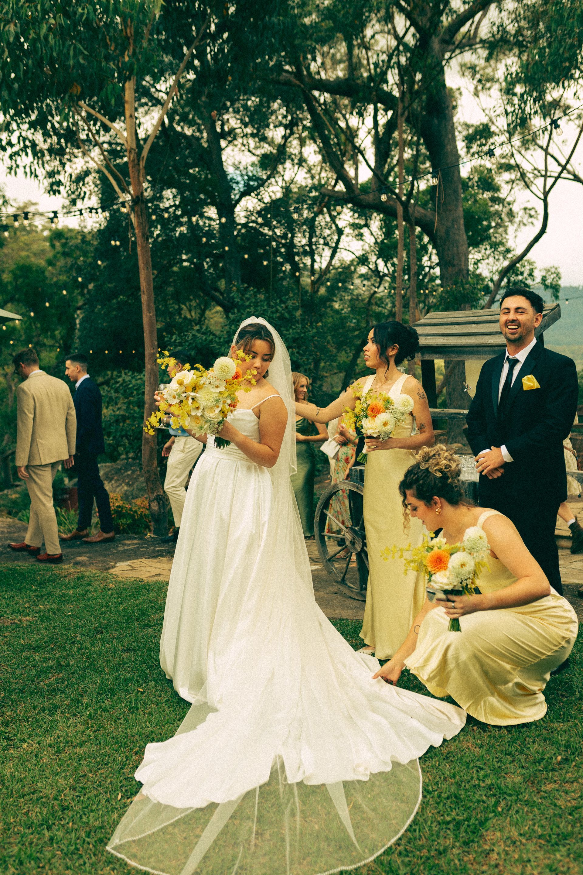 Bride in white dress with bridesmaids and groom at an outdoor wedding; yellow flowers.