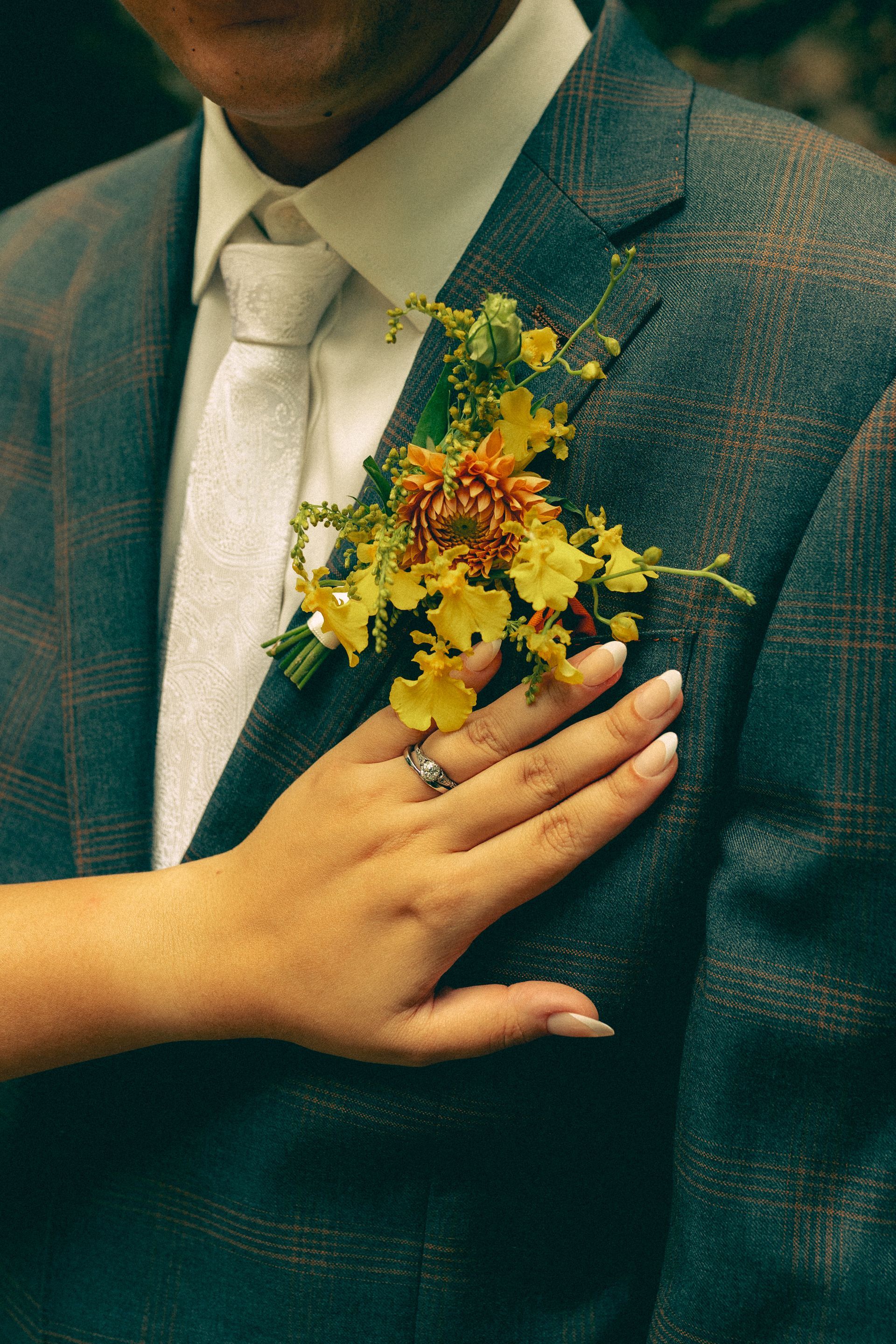 A person in a suit with a flower boutonniere, hand on chest, another hand on top wearing a ring.
