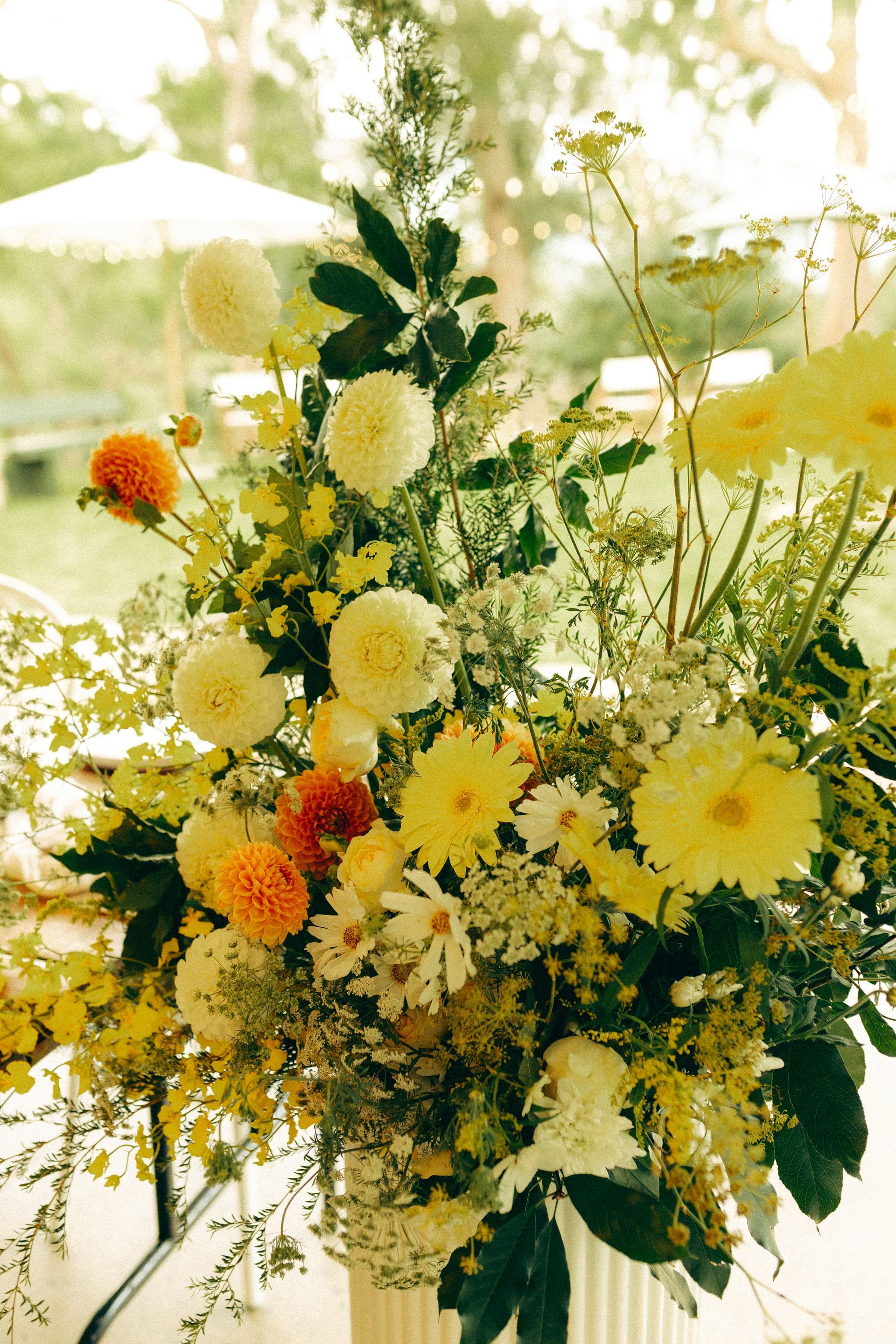 Floral arrangement with yellow and orange flowers, green foliage, in a white vase, outdoors.