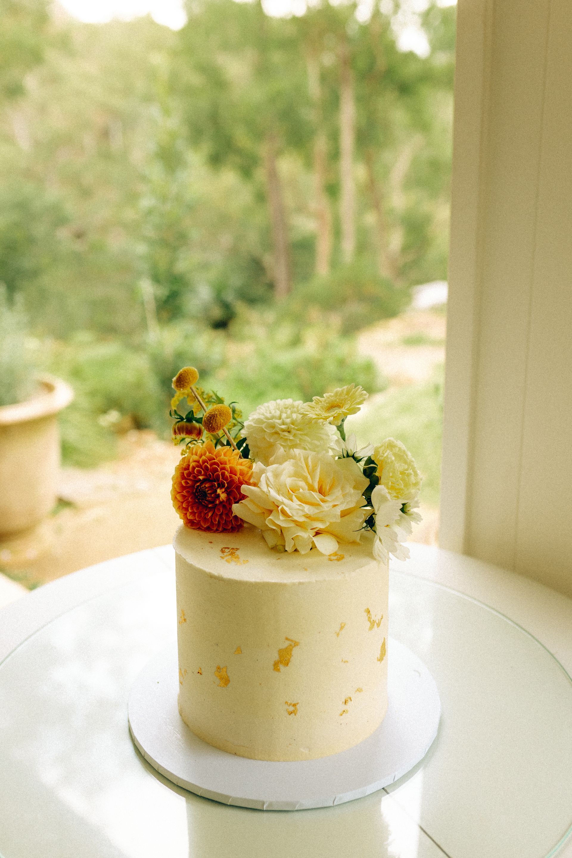 Small cake decorated with flowers and gold flecks on a white plate, set against a blurred green outdoor backdrop.