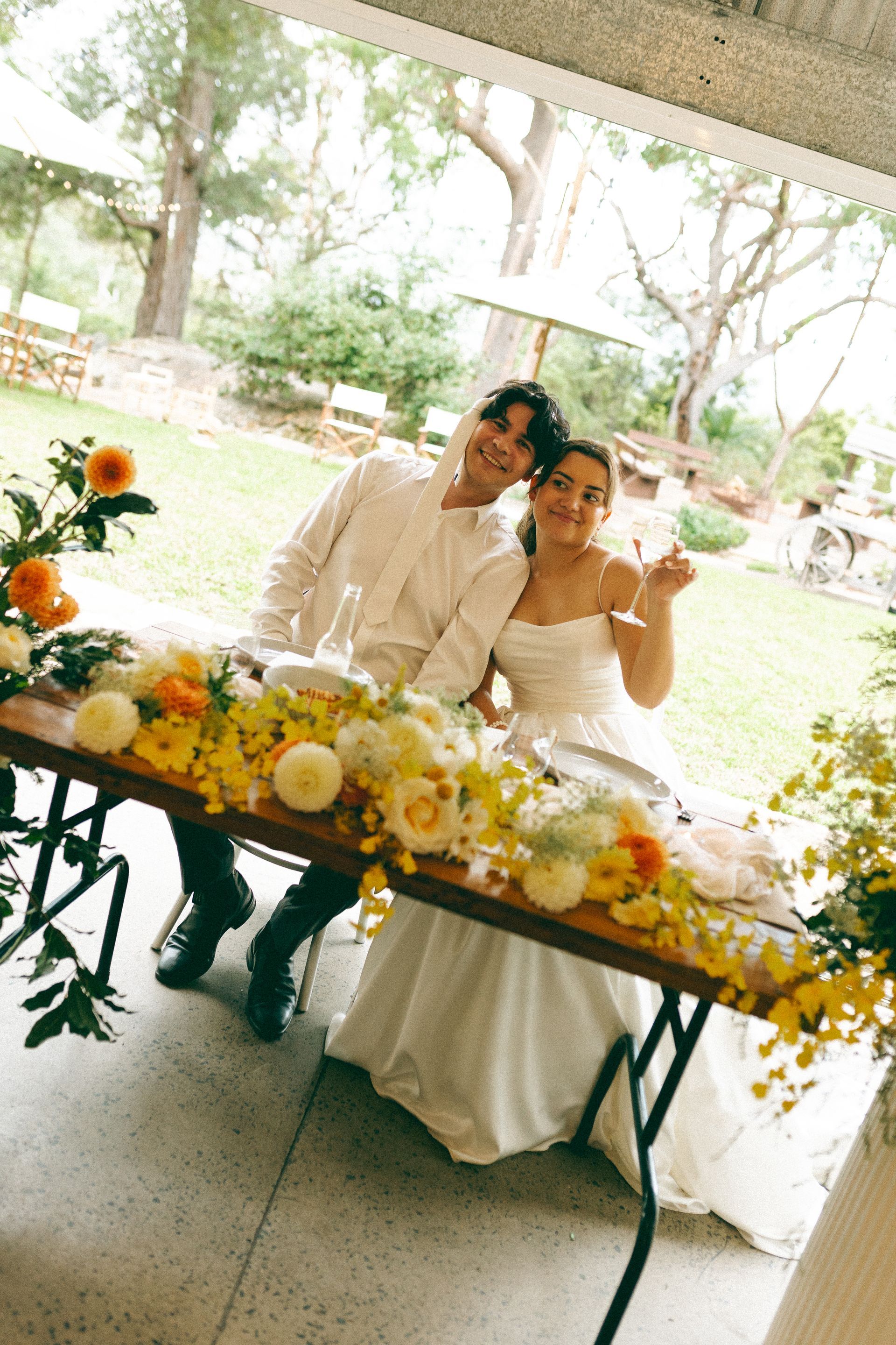 Couple seated at a table decorated with flowers, smiling. Man in white shirt, woman in wedding dress, outdoor setting.