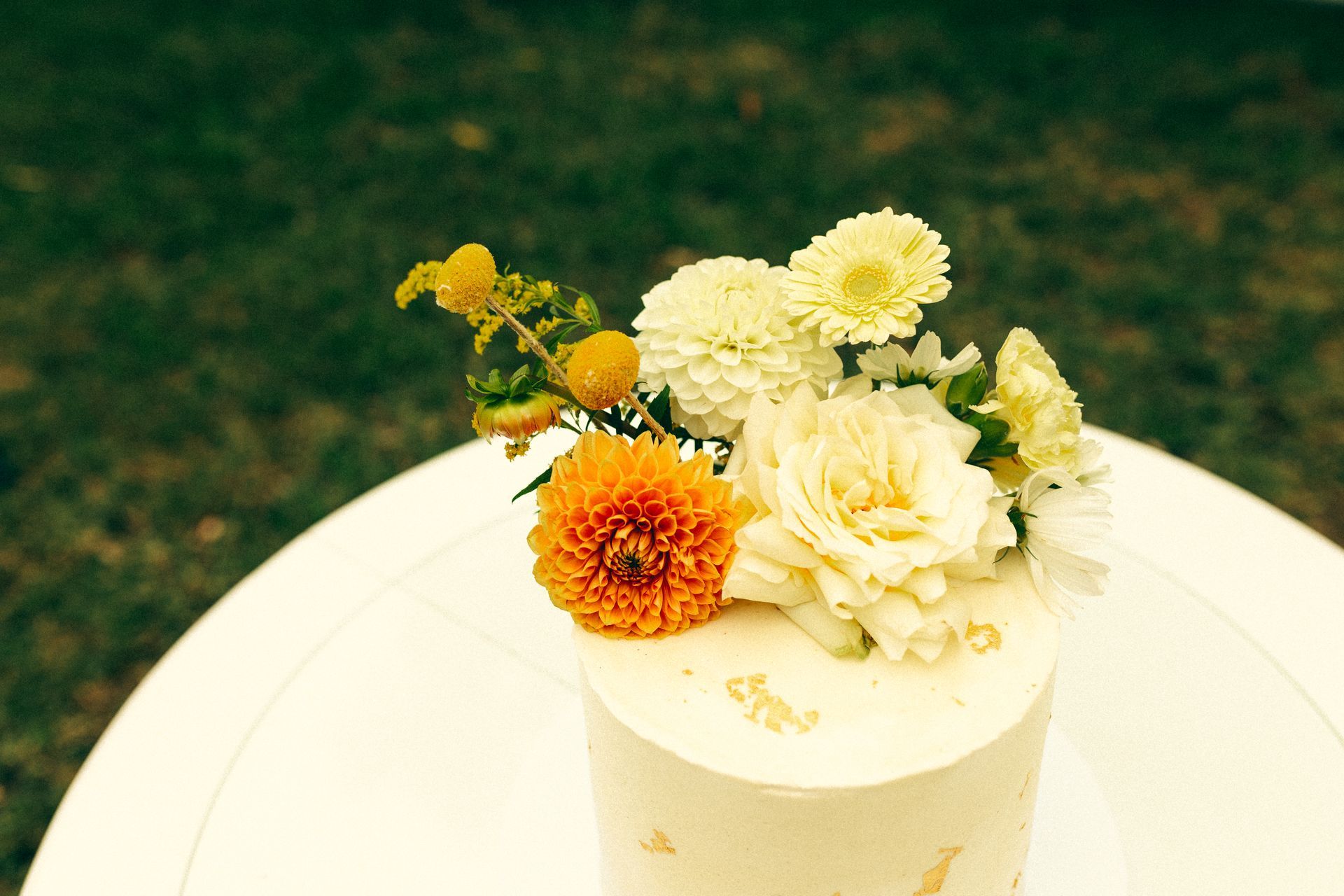 Cake with white frosting and fresh flowers on top, set on a white table with a green lawn background.