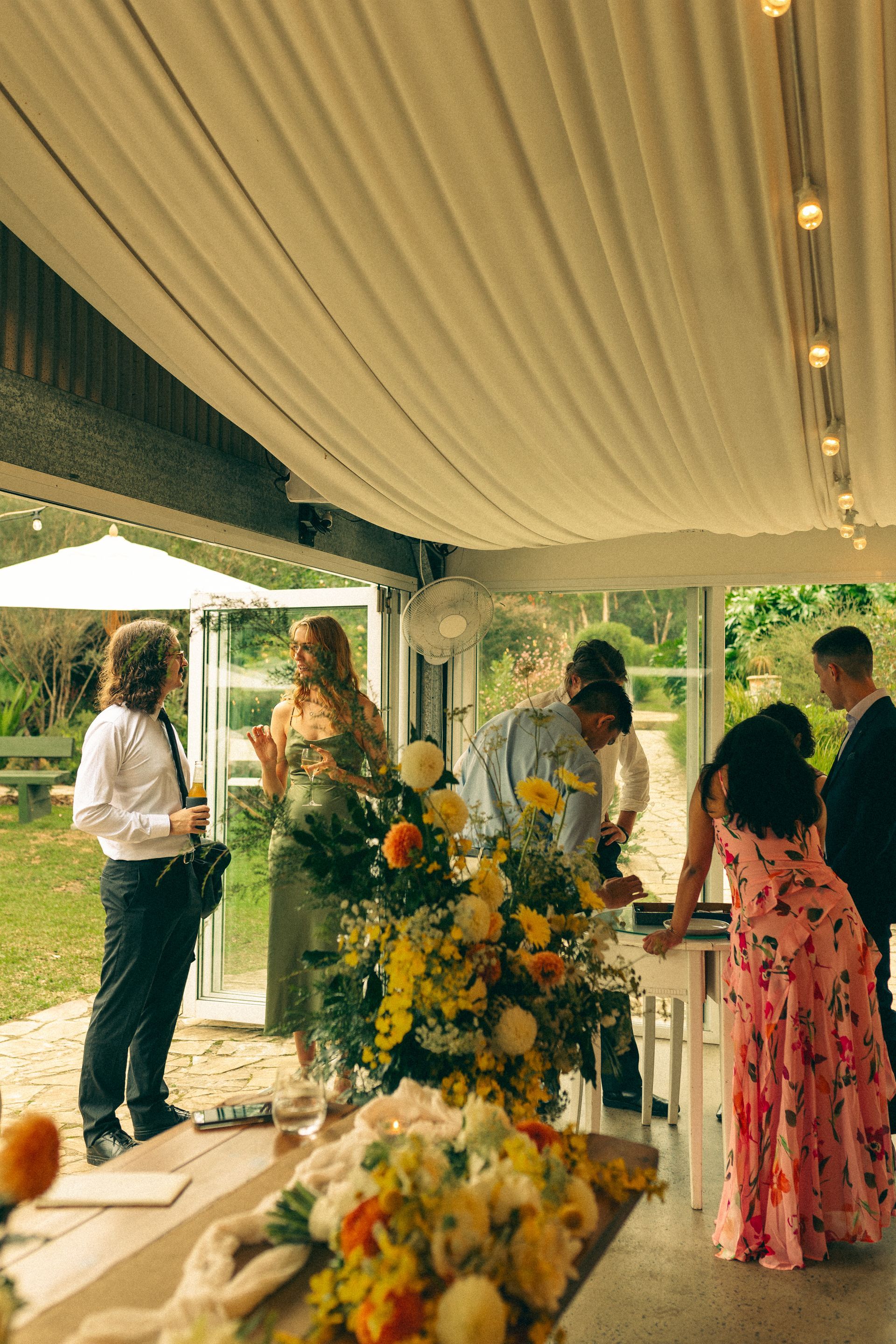 People at an outdoor event, possibly a wedding, near a table with floral arrangements.