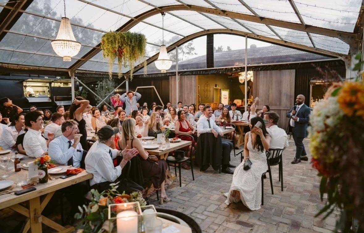 Wedding reception: Bride and groom seated, guests watching, inside a glass-roofed structure with floral décor.
