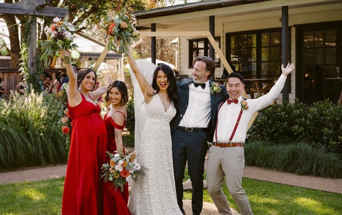 Wedding group cheers with bouquets, bride in white dress, bridesmaids in red, groom in navy, outdoor setting.