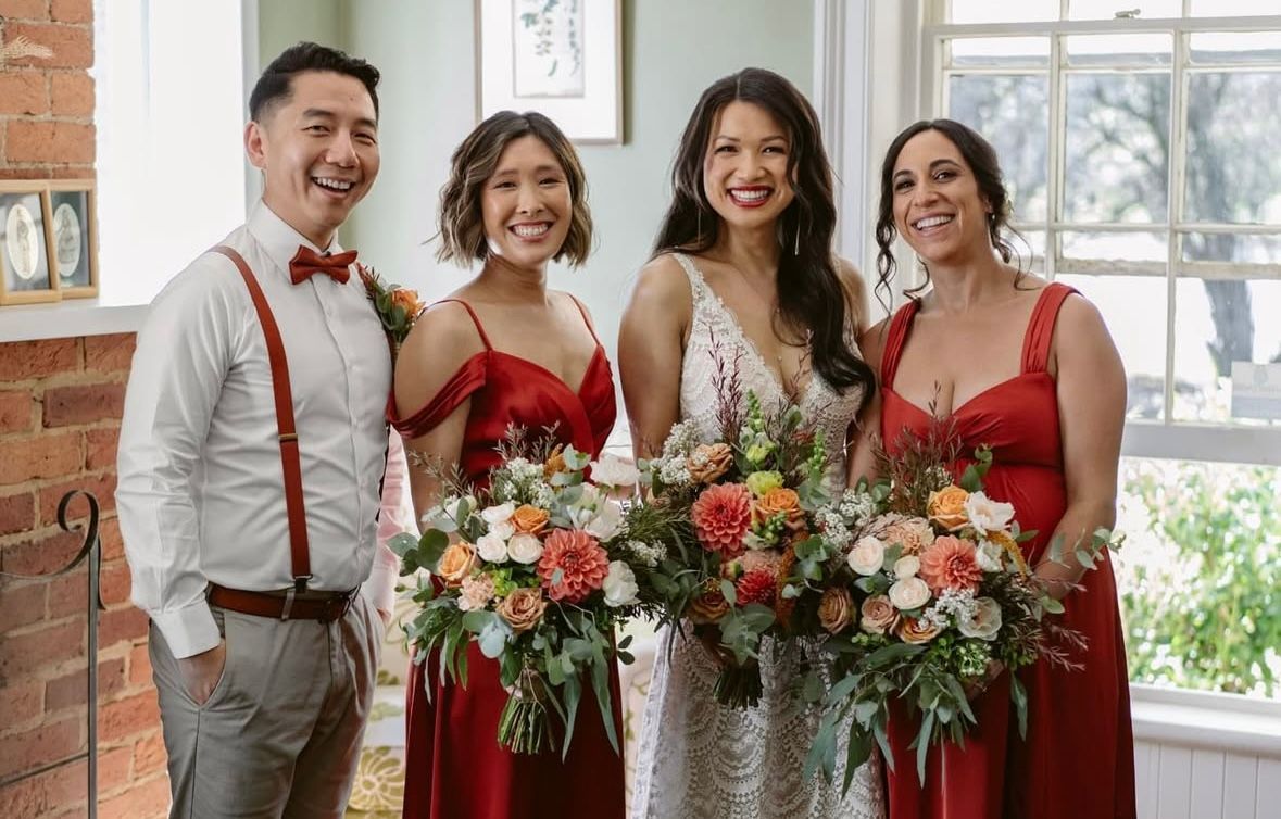Wedding group: Bride with two bridesmaids, a groomsman. All smiling, holding bouquets. Red dresses and bow tie.