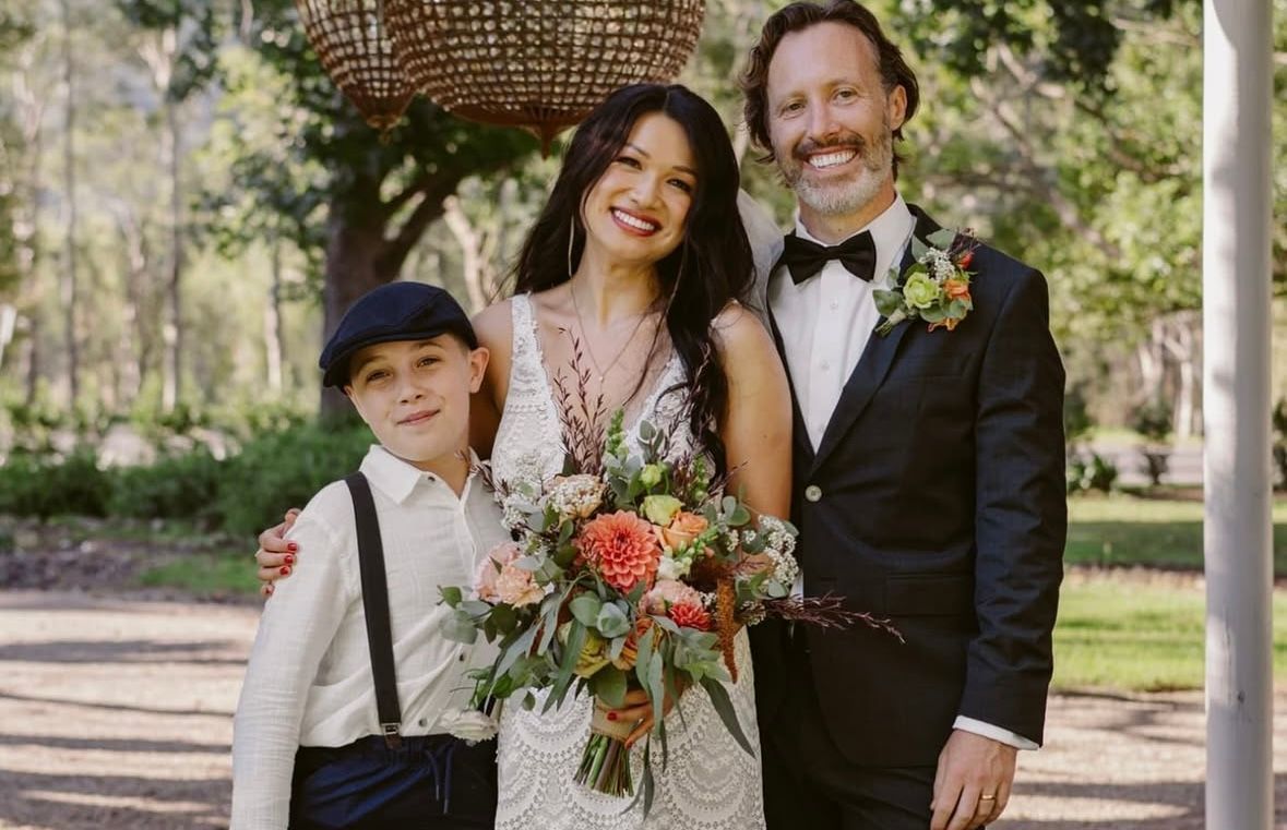 Wedding photo: Bride, groom, and young boy pose outdoors. Bride holds bouquet, smiles; groom in tuxedo.