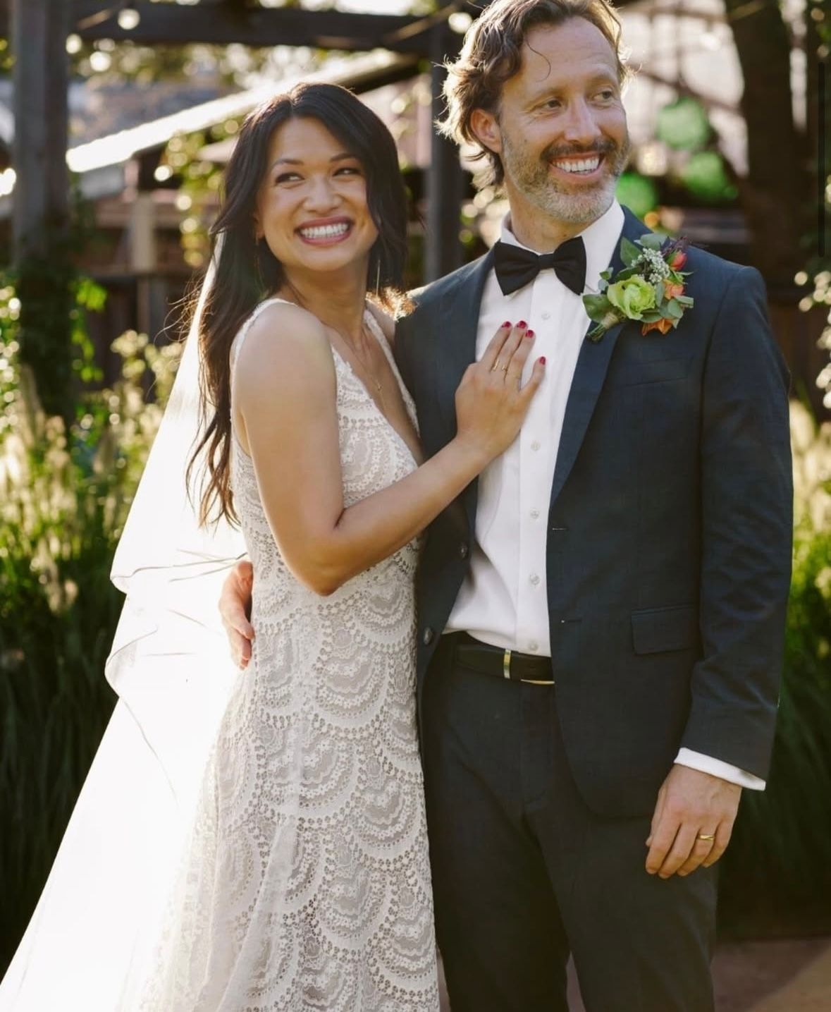Bride and groom smiling, embracing on their wedding day. Woman in lace dress, man in suit with bow tie, outdoors.