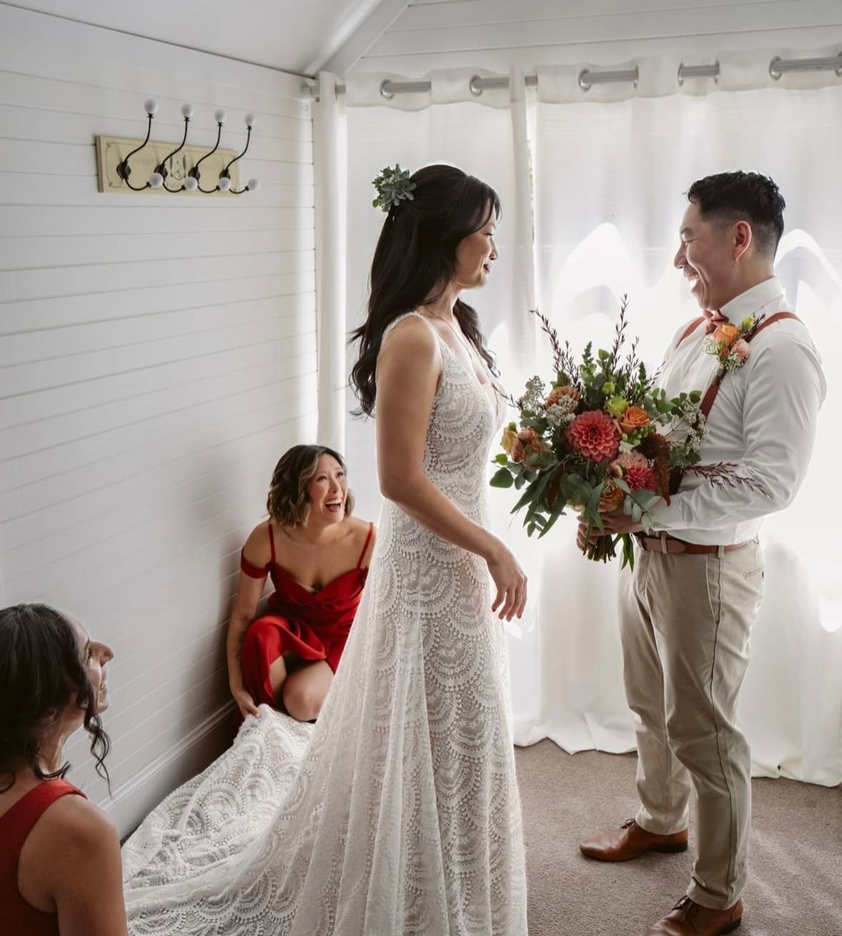 Bride smiles, holding bouquet, as groom reacts. Two bridesmaids in red dresses assist in a white room.