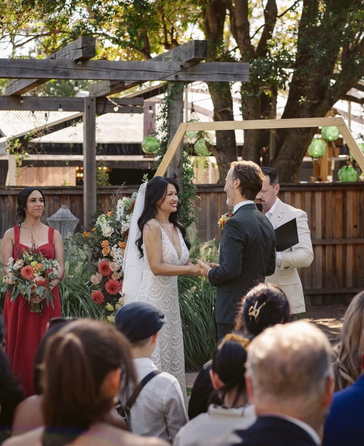 Wedding ceremony: Bride and groom exchanging vows, guests watching outdoors under arbor.