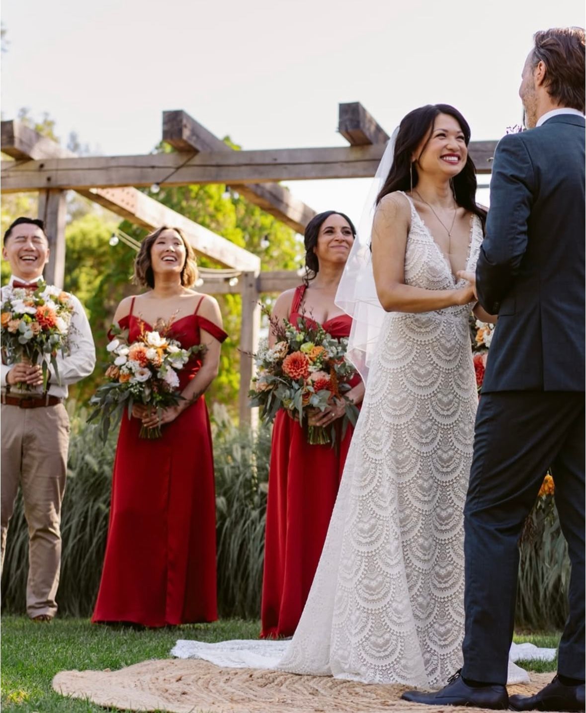 Bride smiling during outdoor wedding ceremony; bridesmaids in red dresses; groom in green suit.