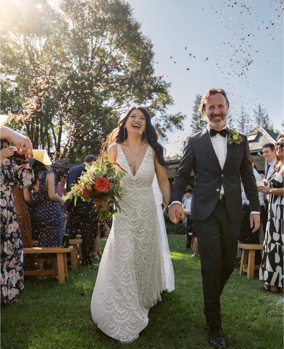 Bride and groom walk down aisle, smiling. Confetti falls. Outdoors, sunny wedding.