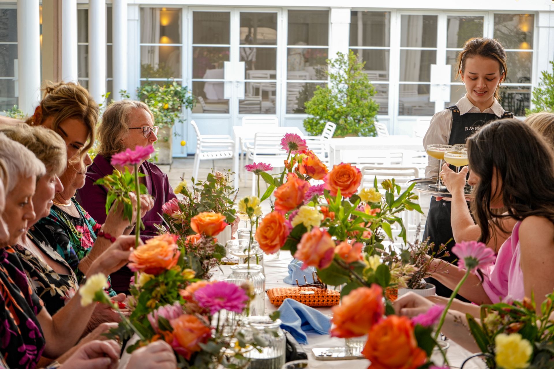 Women at a floral arranging workshop. Bright flowers on a table. Server brings drinks. White exterior.