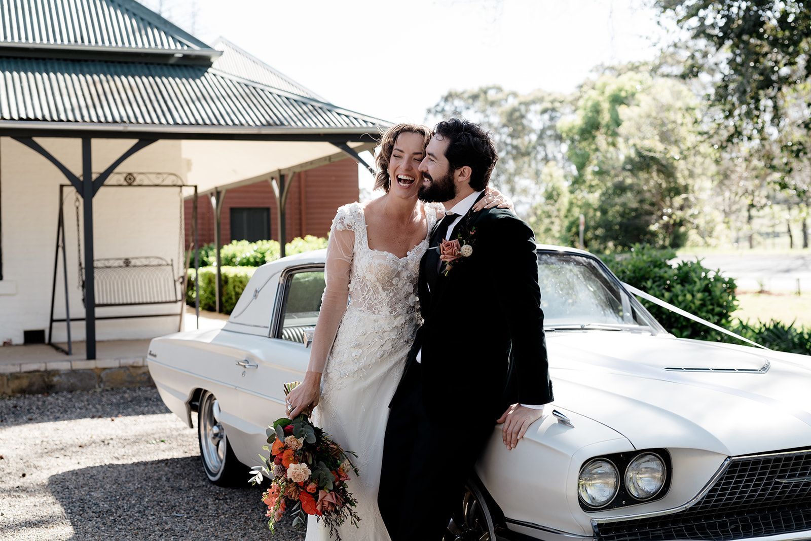 Newlyweds laugh, leaning on a white vintage car outside a building with a veranda.
