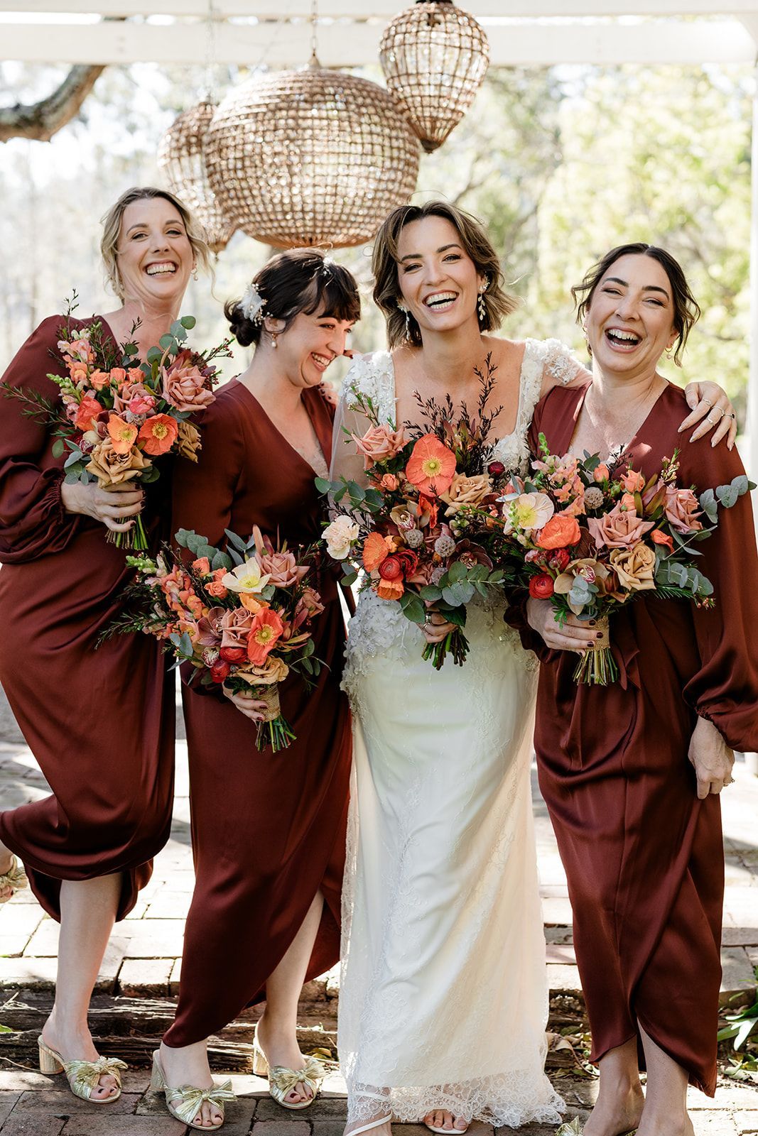Bride and bridesmaids laughing, holding bouquets, wearing maroon dresses, and standing outdoors.
