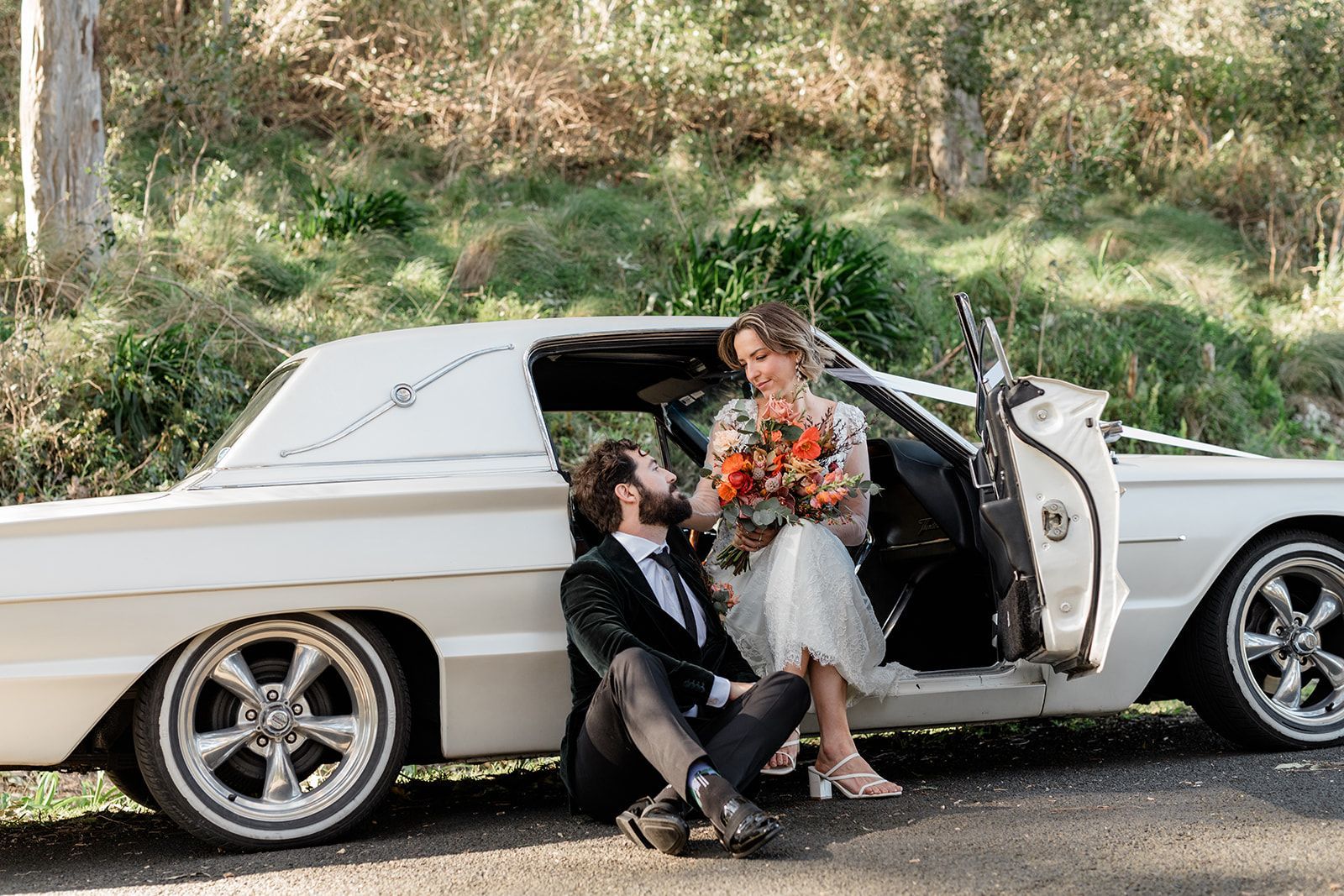 Newlyweds pose by a white classic car; woman in floral dress, man in a suit, both smiling, outdoors.