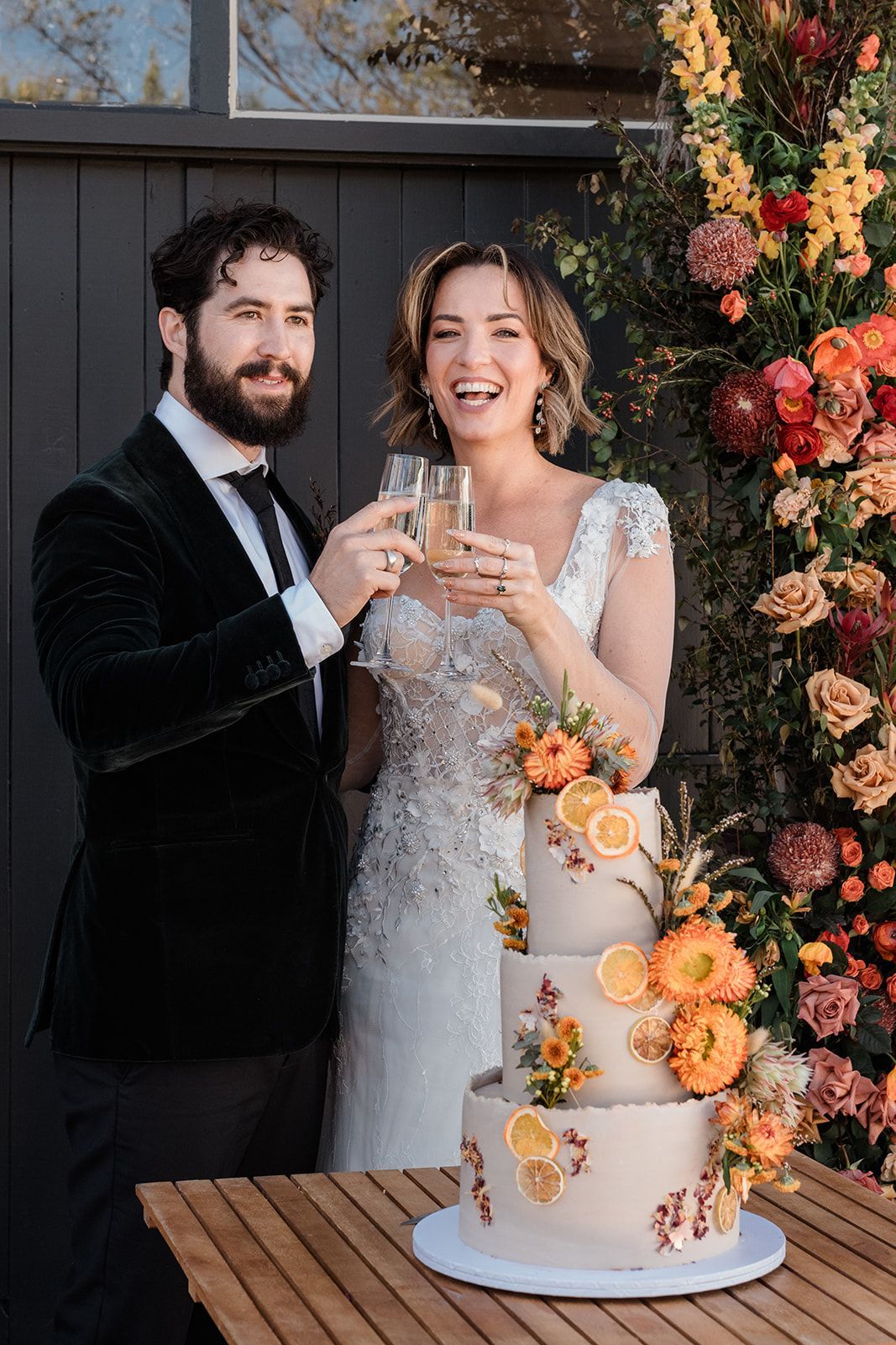 Couple toasting champagne next to a decorated wedding cake. Orange, yellow floral archway in the background.