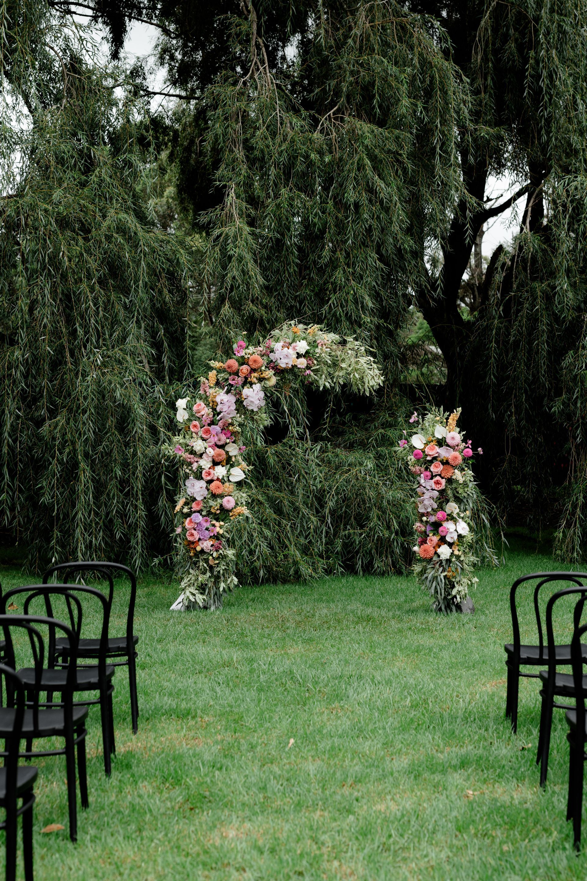 Floral wedding arch and columns in a grassy outdoor setting with black chairs.