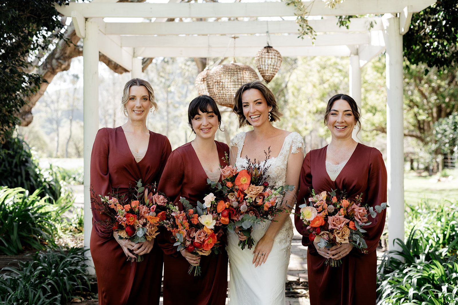Bride and bridesmaids in burgundy dresses holding bouquets, pose under an arbor.