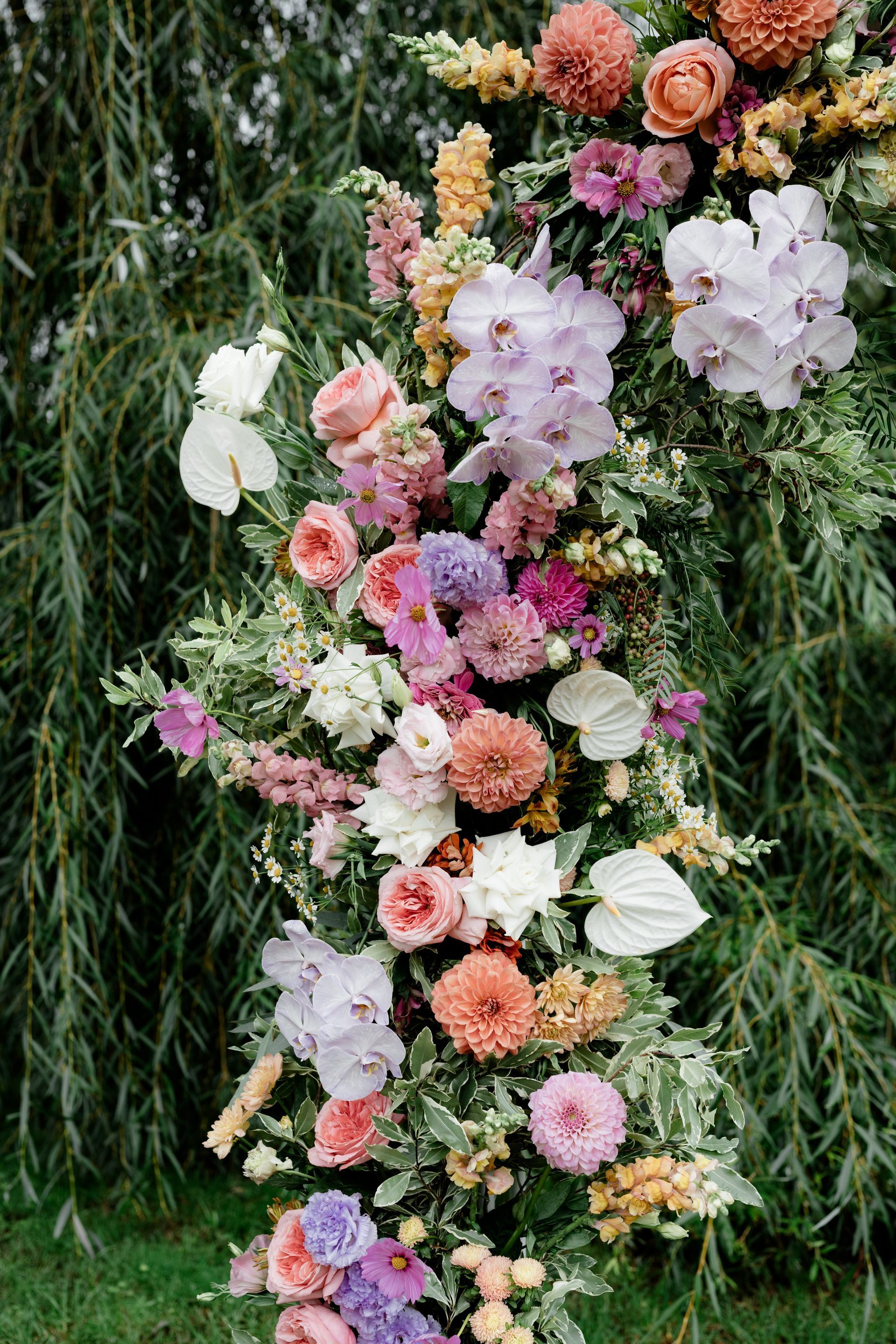 Floral arch with orchids, roses, and dahlias in pinks, oranges, and purples against a green backdrop.