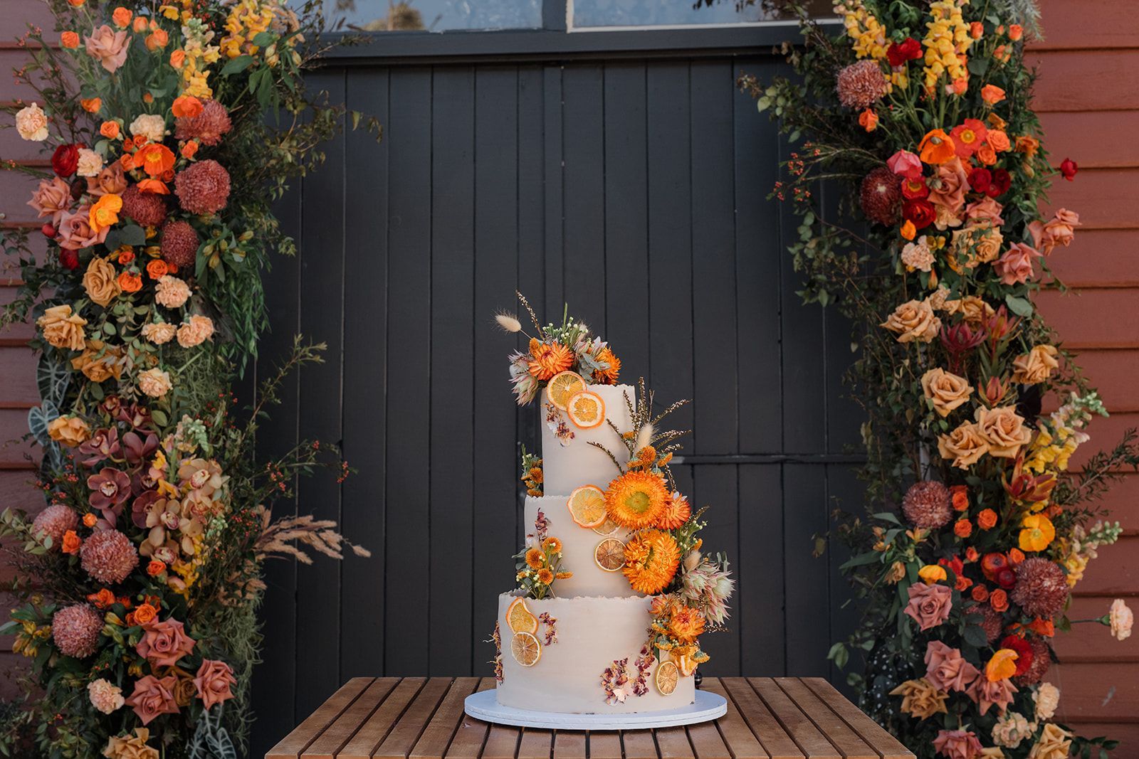 Three-tiered wedding cake decorated with flowers and orange slices, set against a floral arch and gray door.