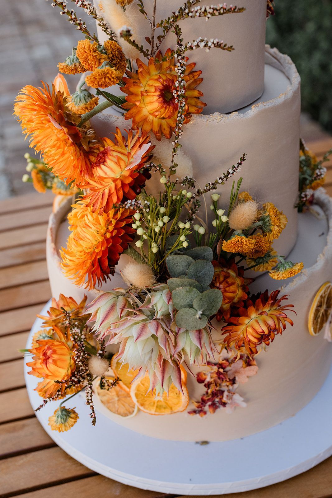 Three-tiered wedding cake, tan frosting, decorated with orange and yellow flowers and dried orange slices.
