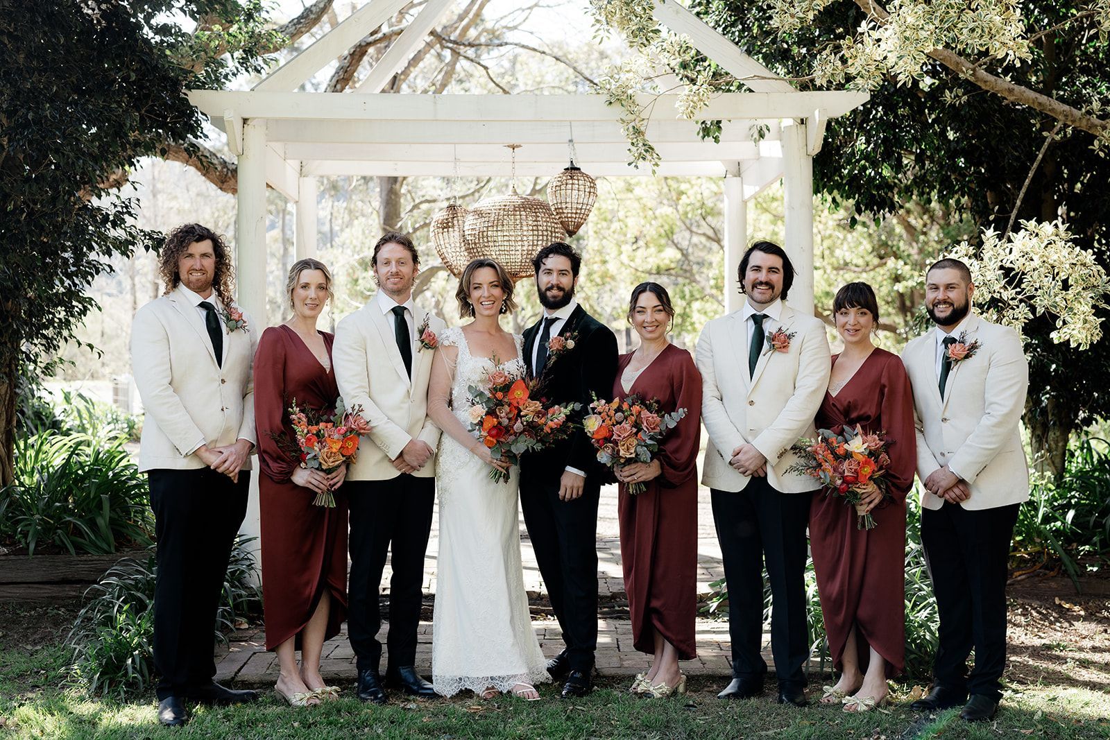Wedding party posing in front of white arbor. Bride and groom centered; bridesmaids in burgundy, groomsmen in cream suits.