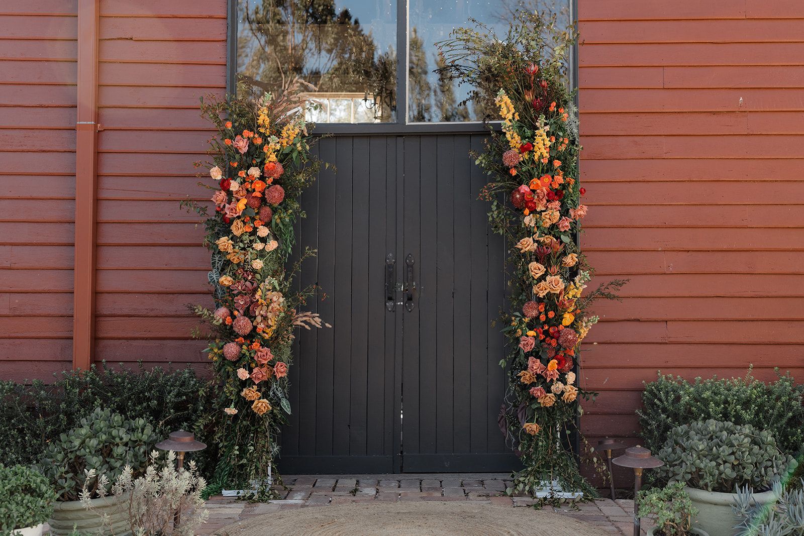 Floral arrangements frame a dark wooden door on a red building.