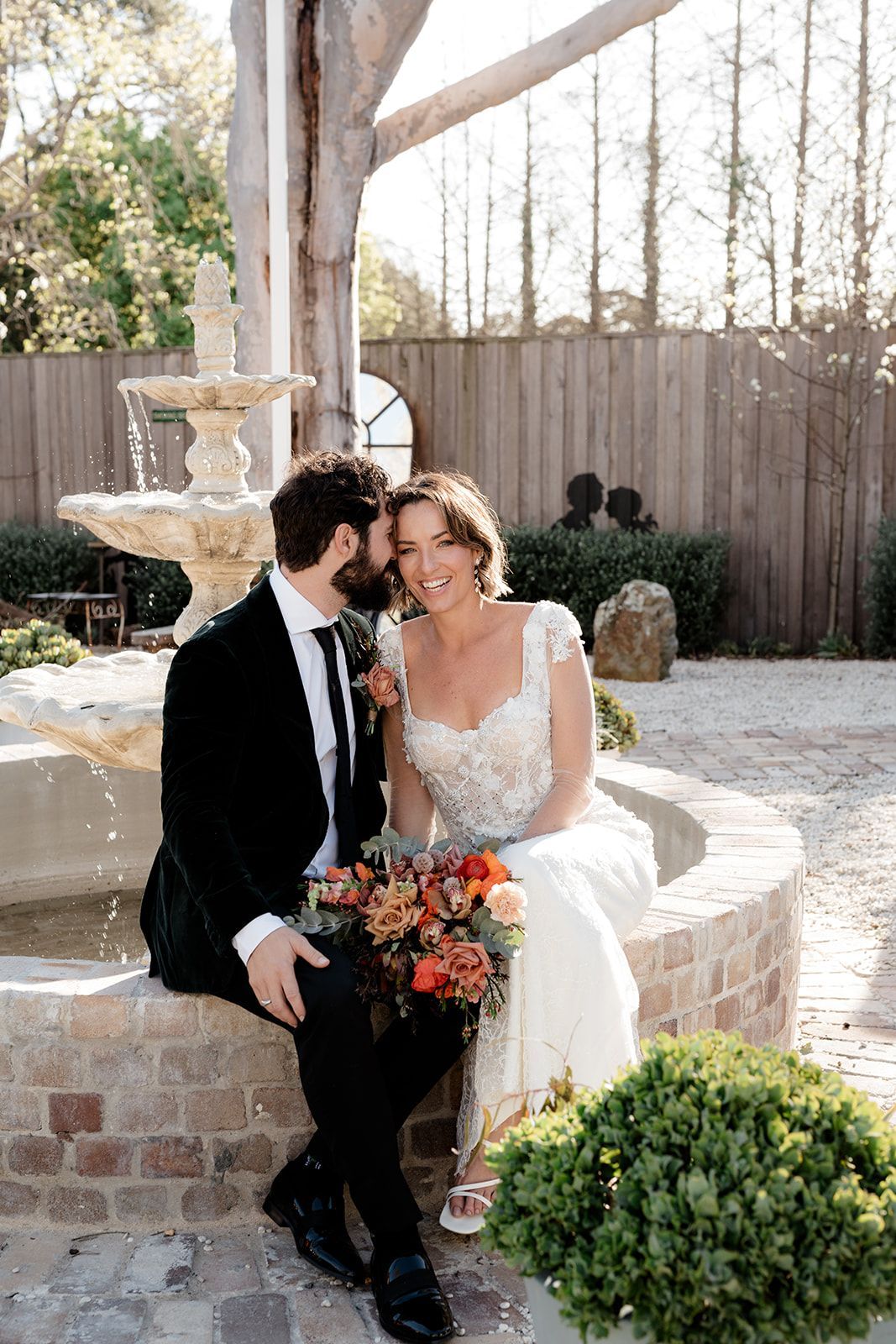 Bride and groom sit by a fountain, smiling. The groom whispers in her ear. They hold a bouquet.