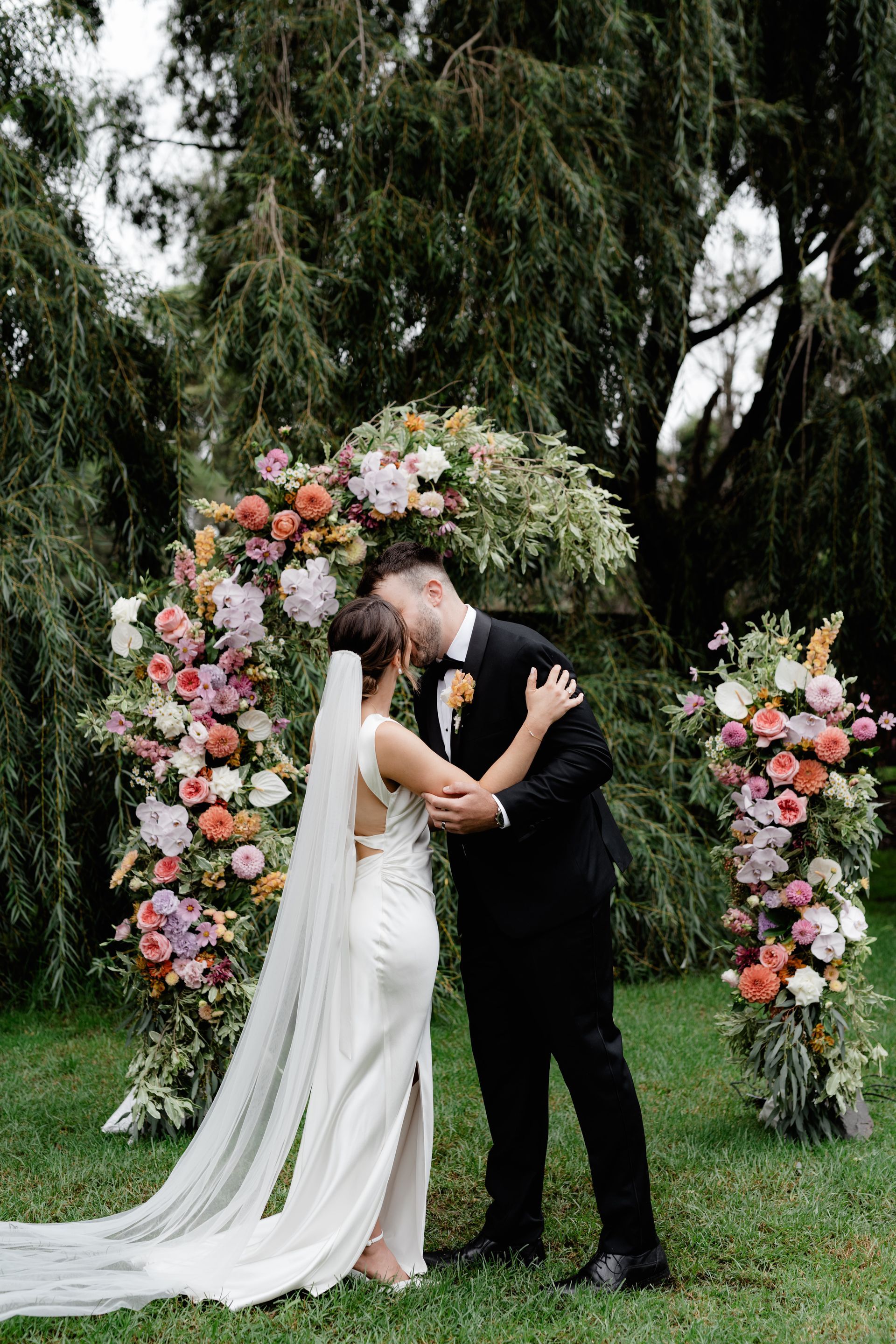 Bride and groom kissing under a floral archway at outdoor wedding.