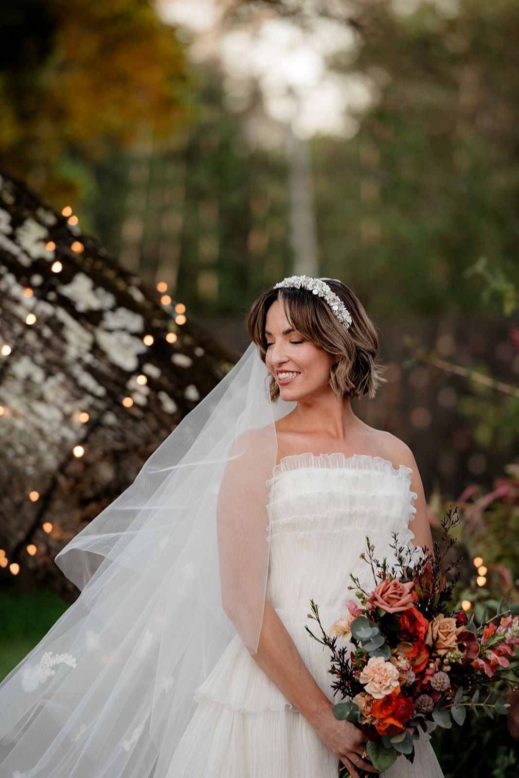 Bride in white dress, holding bouquet, smiling, veil, beaded headband, outdoors, fall foliage, string lights.