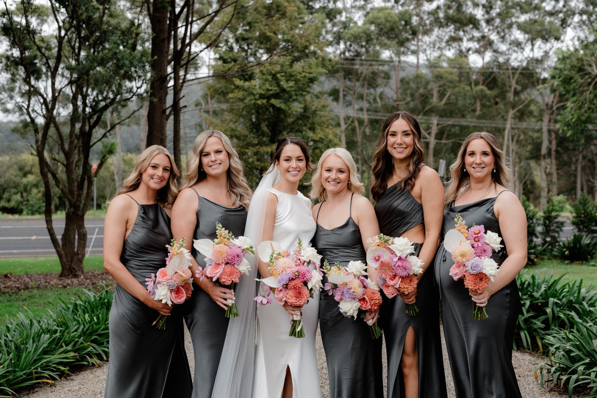 Bride in white gown with bridesmaids in gray dresses holding bouquets, posing outdoors.