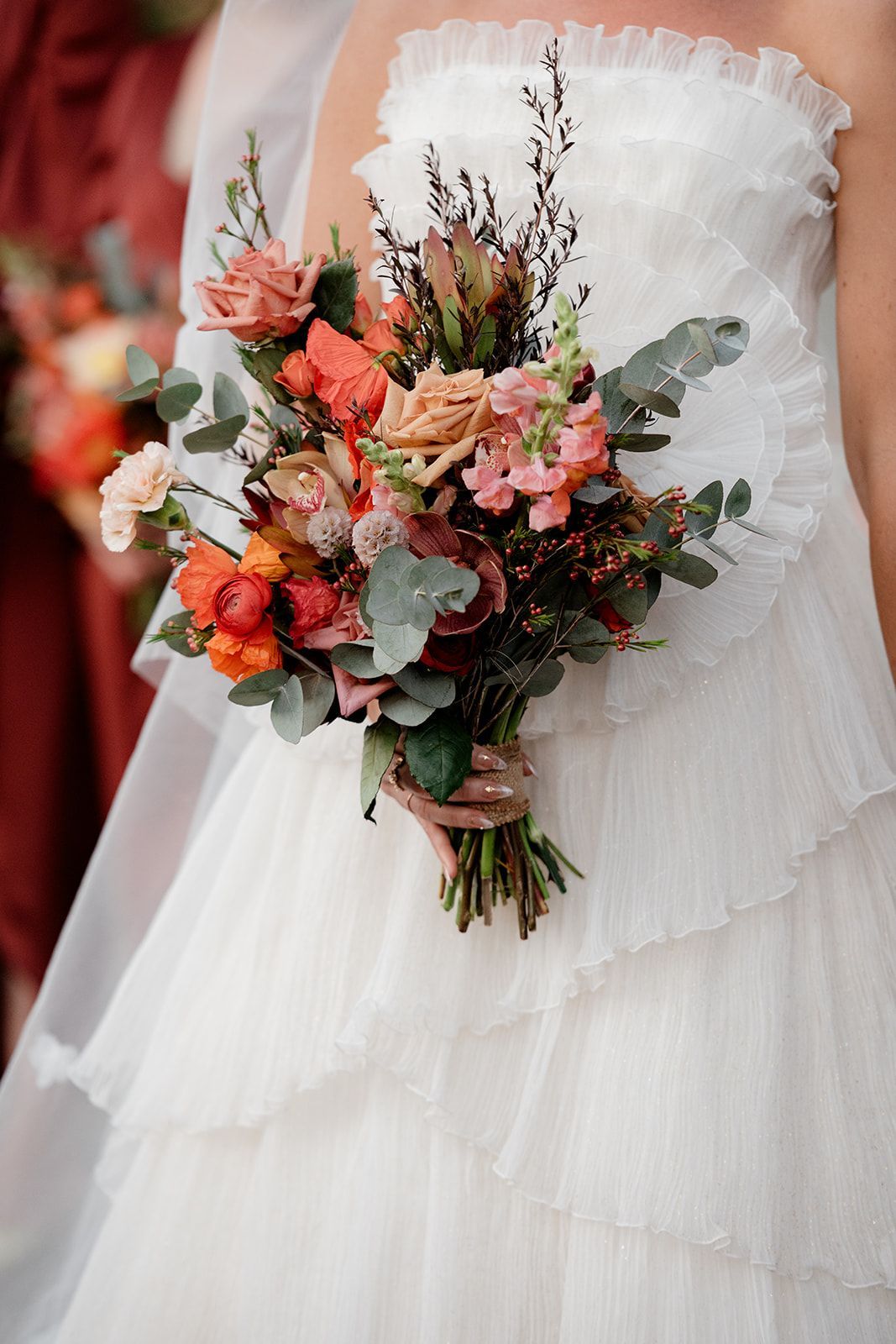 Bride in a white tiered dress, holding a bouquet of orange and pink flowers, with greenery.