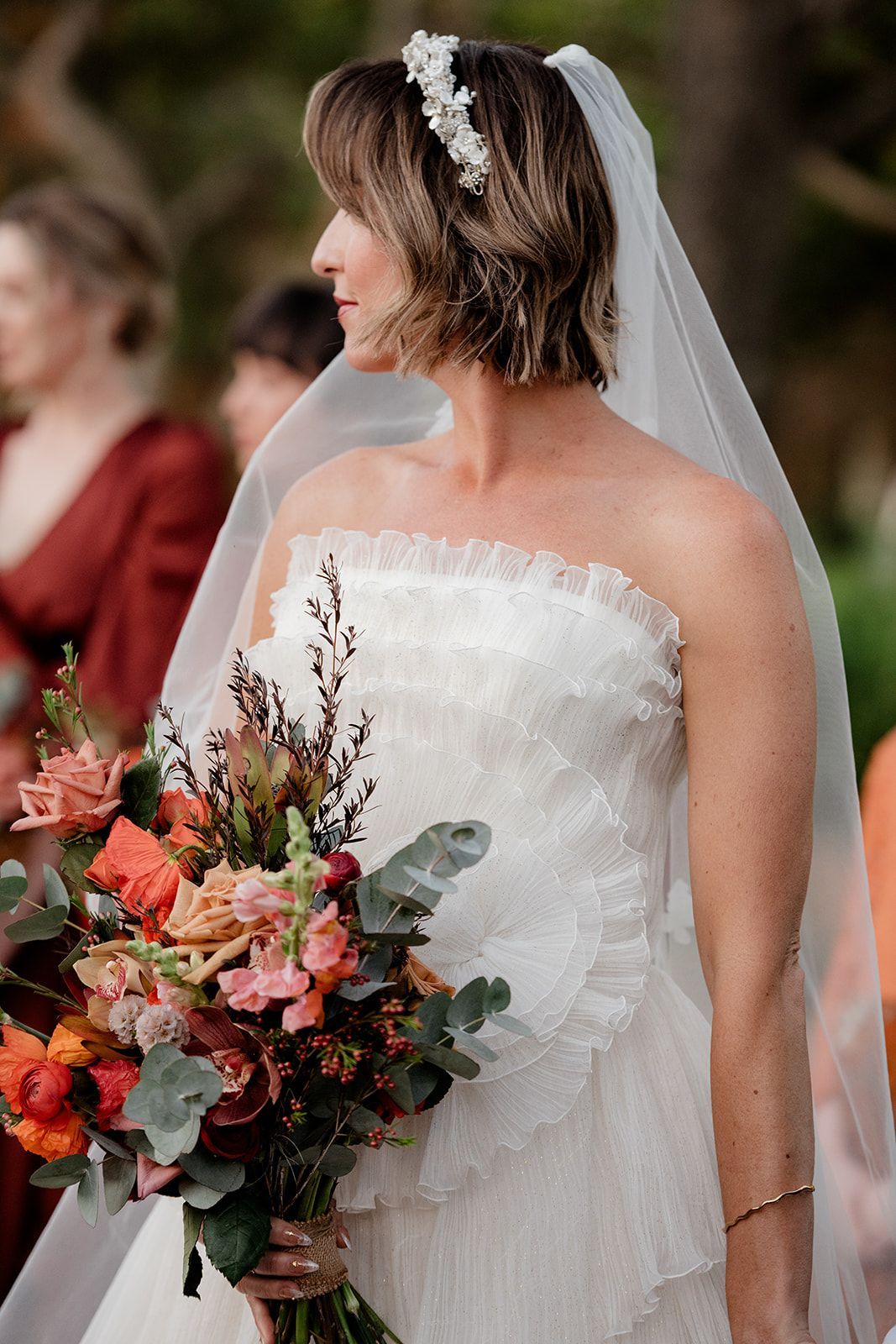 Bride in a strapless white lace dress and veil, holding a bouquet, standing outside.