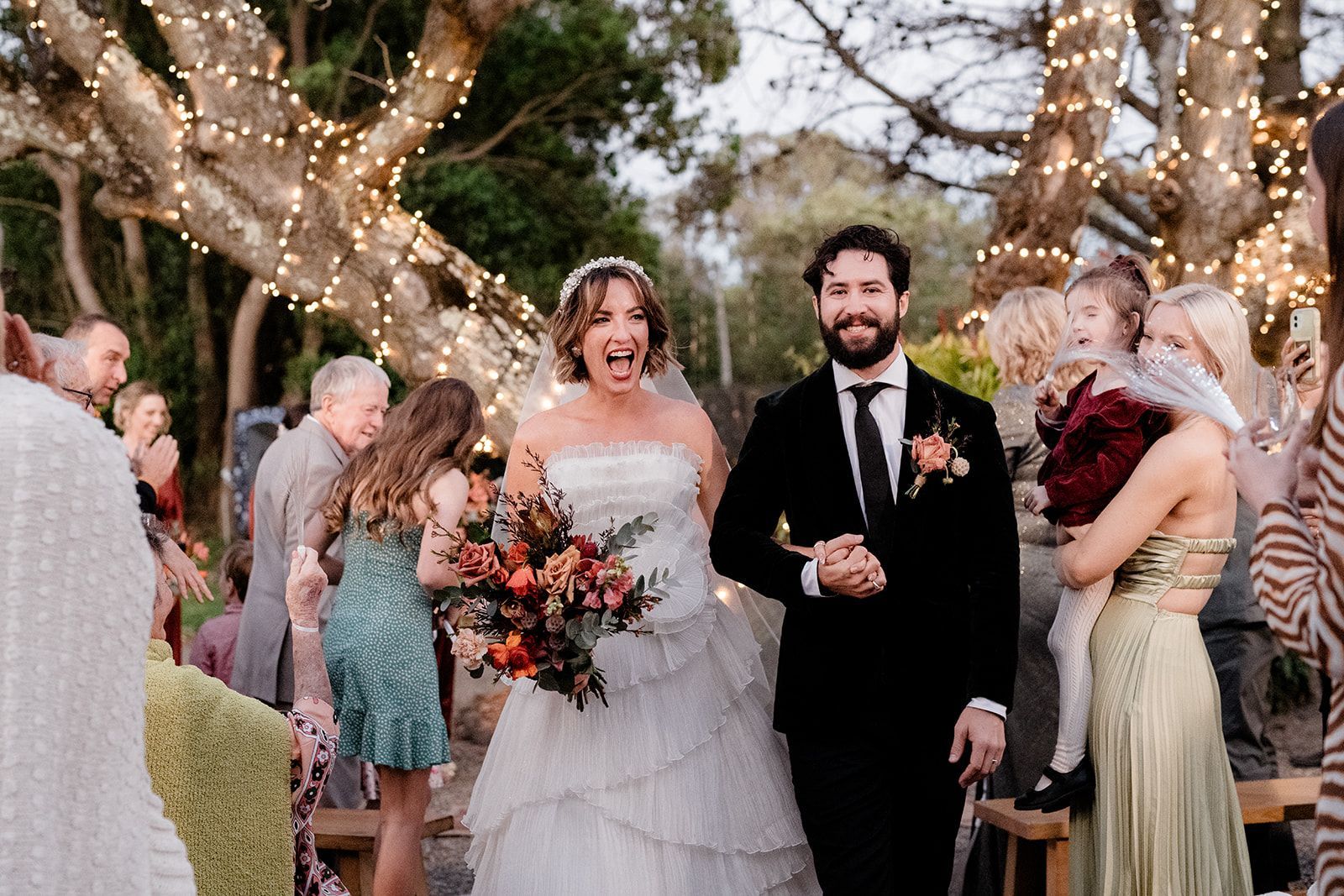 Newlyweds walking, bride smiling wide, groom beside her, guests cheering, trees with lights.