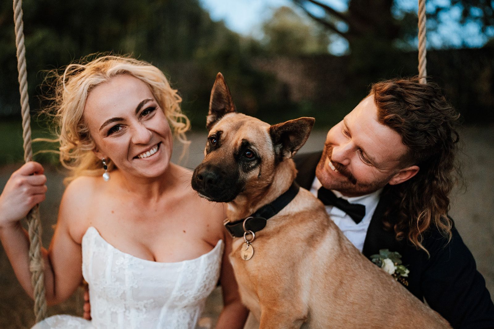 Bride and groom smile with their dog on a swing set. She wears a white dress; he has a black suit.