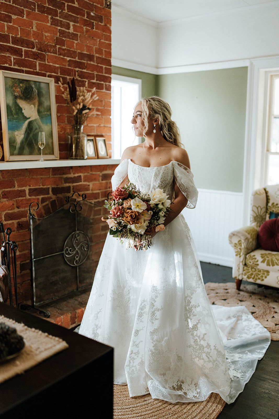 Bride in a white lace gown holding bouquet, standing near a brick fireplace, looking out a window.