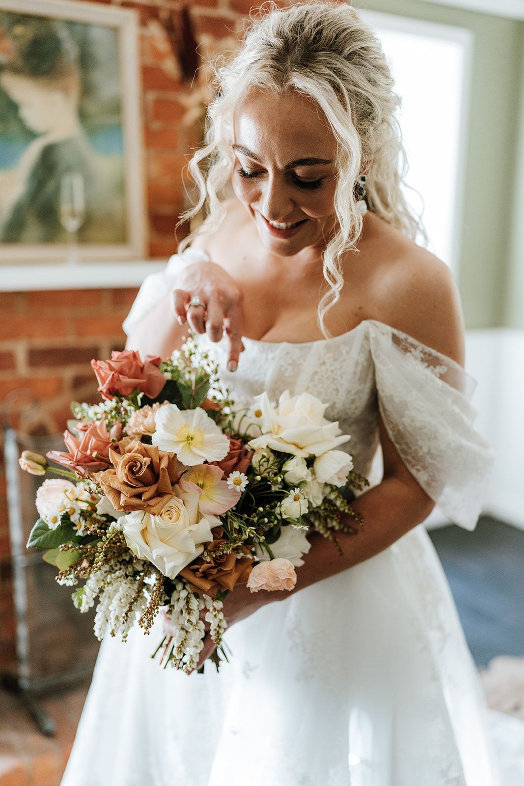 Bride in a white off-the-shoulder dress smiles at her wedding bouquet, set against brick and floral decor.