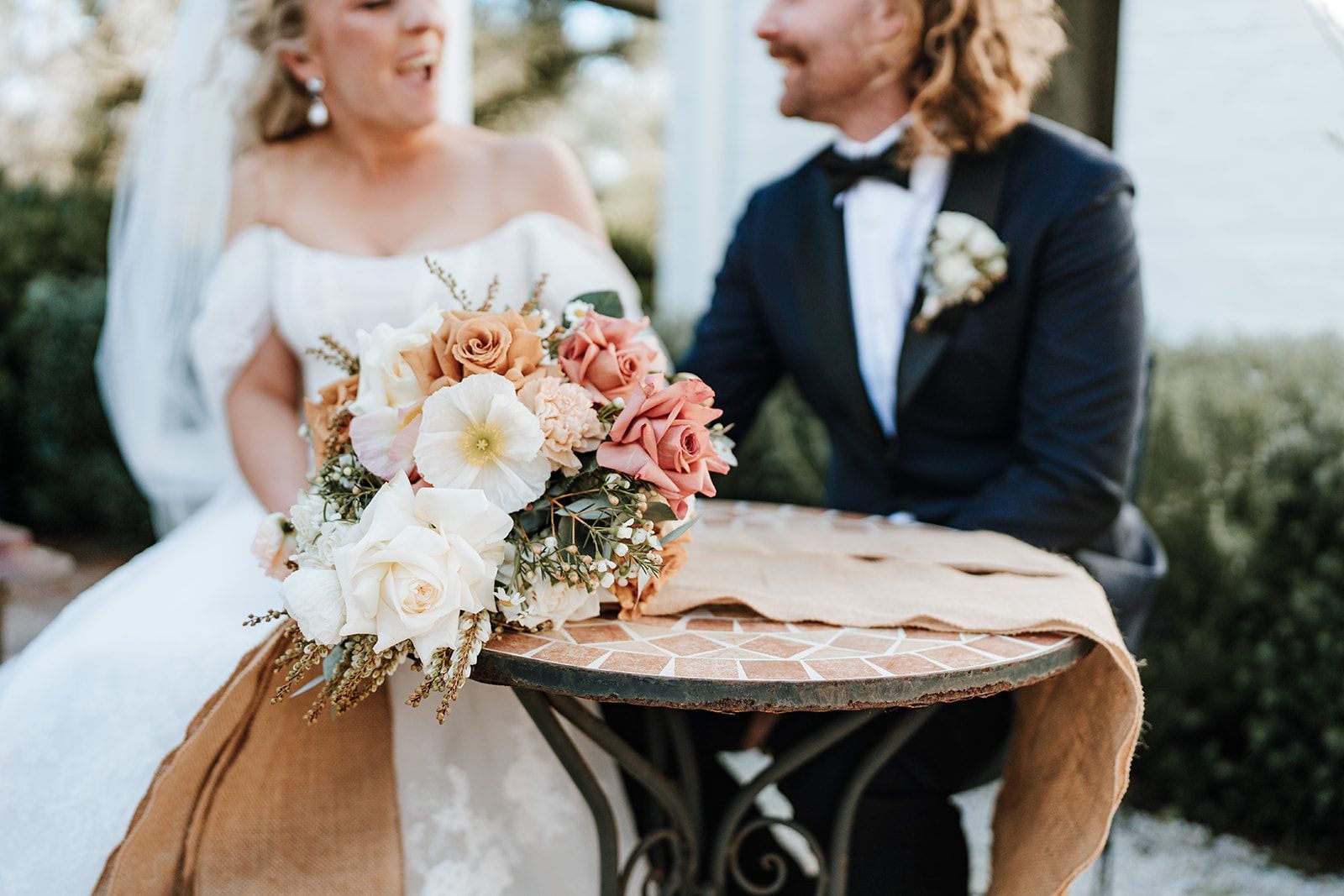 Bride and groom laughing, flowers on a table. Exterior shot, sunny day.