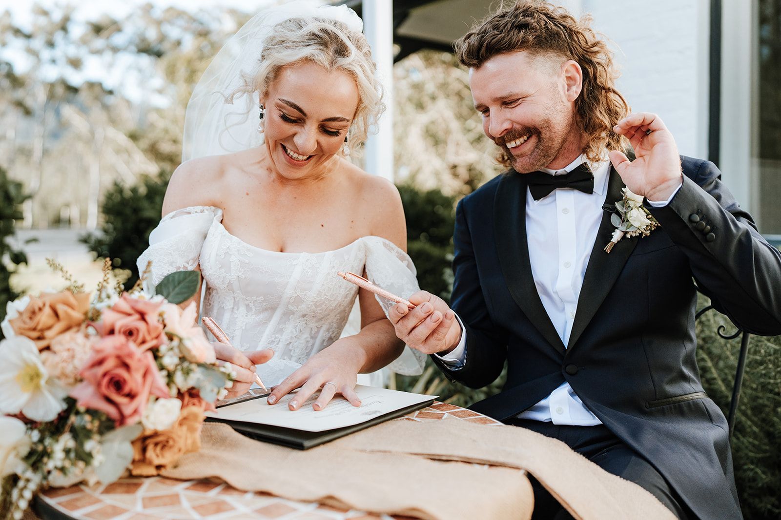 Bride and groom sign a document at a table, smiling. She wears a wedding dress; he wears a tuxedo.