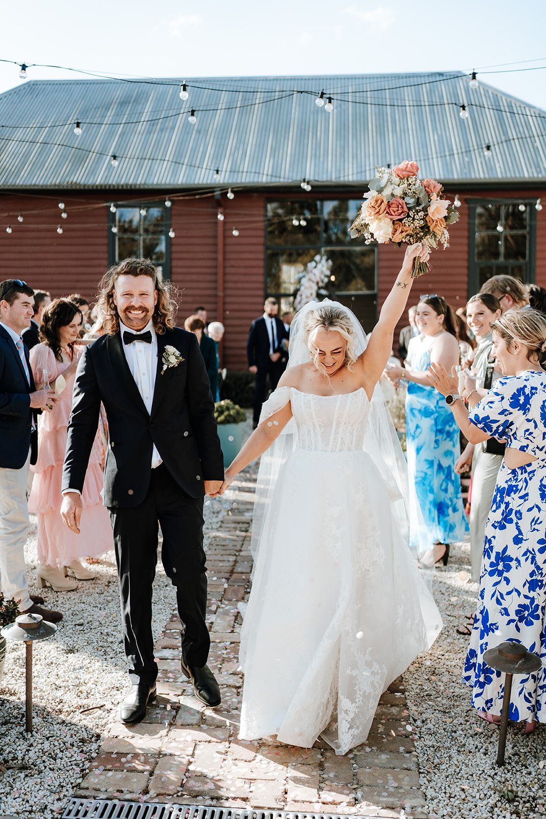 Newlyweds walking, holding hands, celebrating with guests near a brick building.