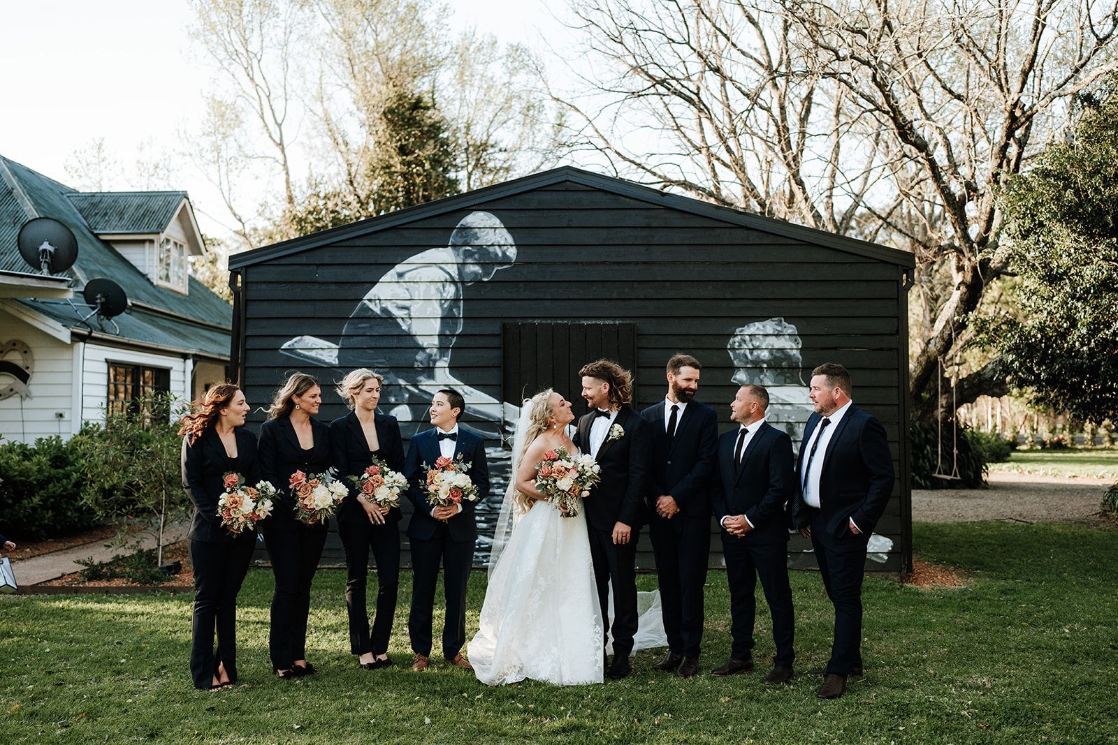 Wedding party poses in front of a black building with a mural. Bride and groom centered; others in dark suits.