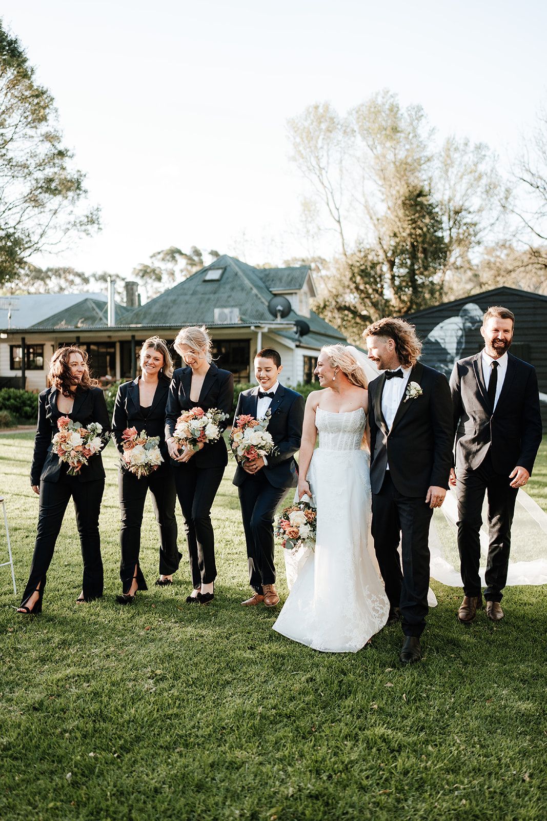 Wedding party walking on grass: bride in gown, groom in suit, bridesmaids in pantsuits, sunny setting.