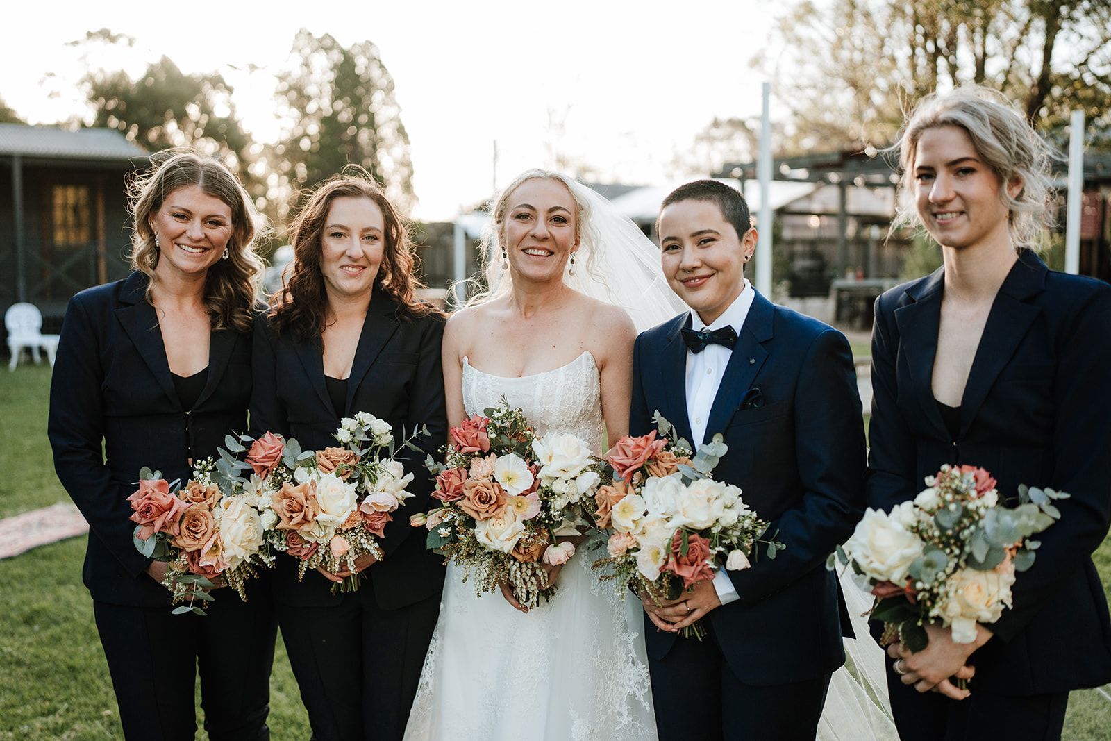 Bride and bridesmaids in suits and dresses holding bouquets, outdoor wedding.