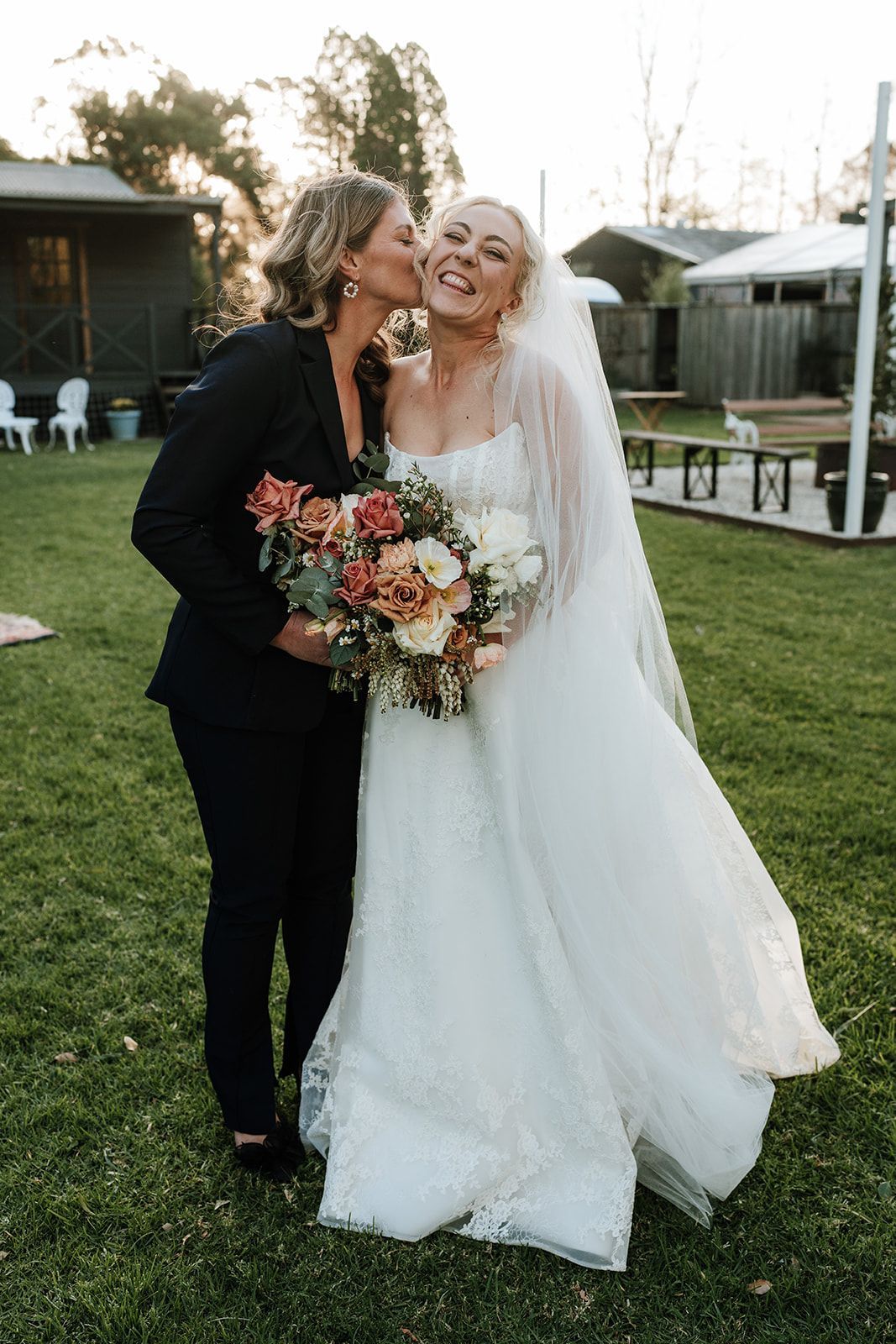 Woman in tuxedo kisses a bride in a wedding dress, holding flowers, outdoors.